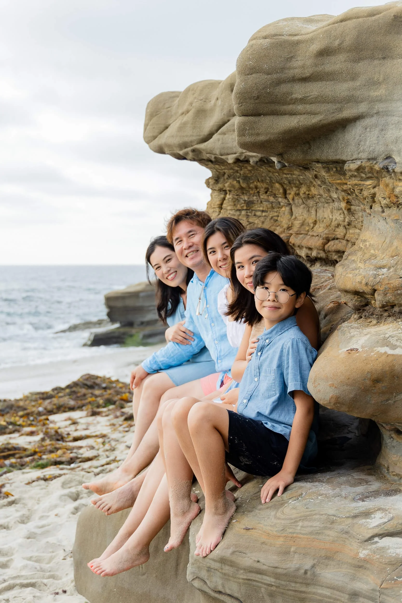 A group of five smiling people sitting on a rock ledge at the beach, with the ocean and cloudy sky in the background.