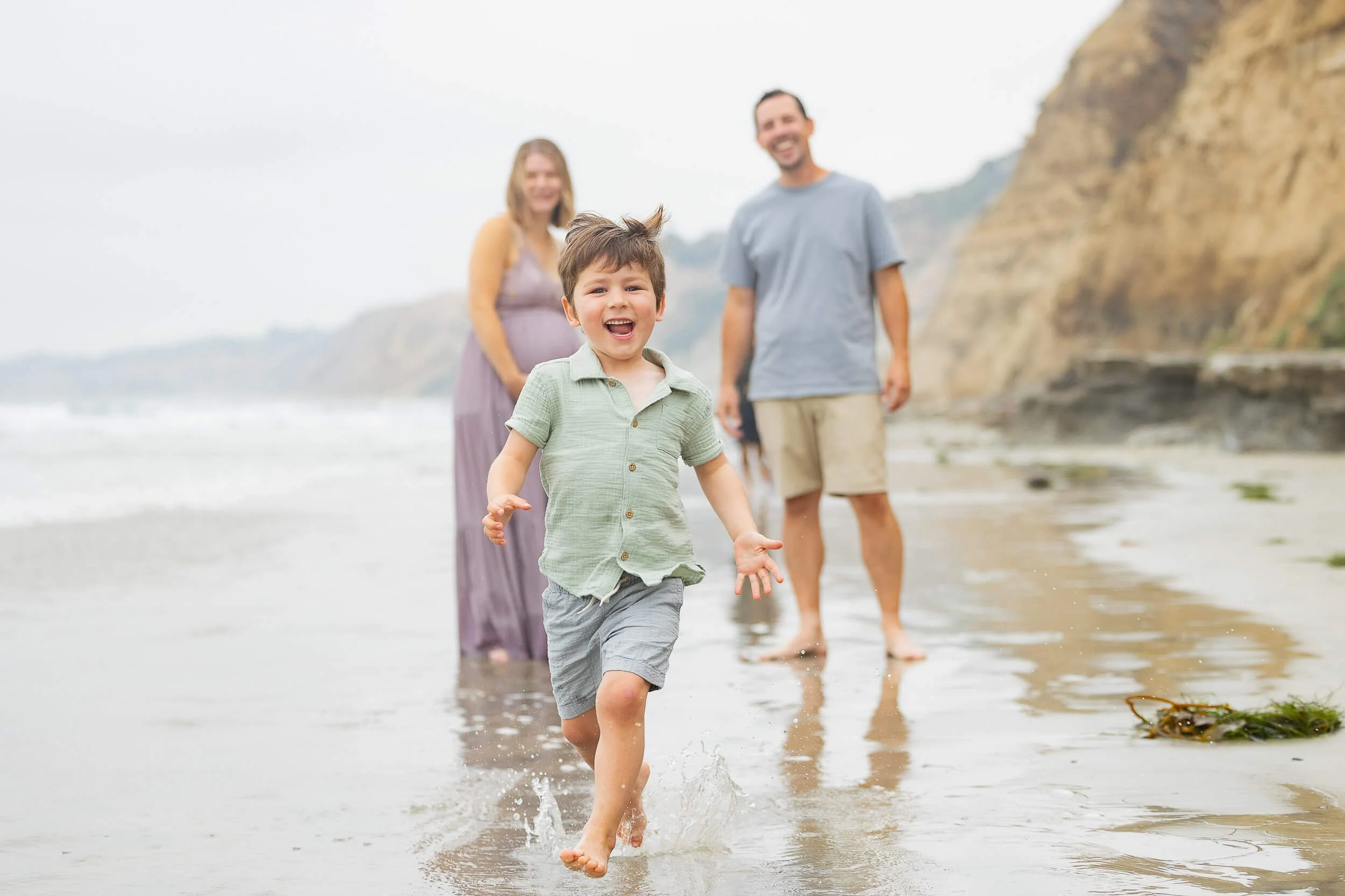 A young boy running and smiling on a beach with family members walking behind him near cliffs.