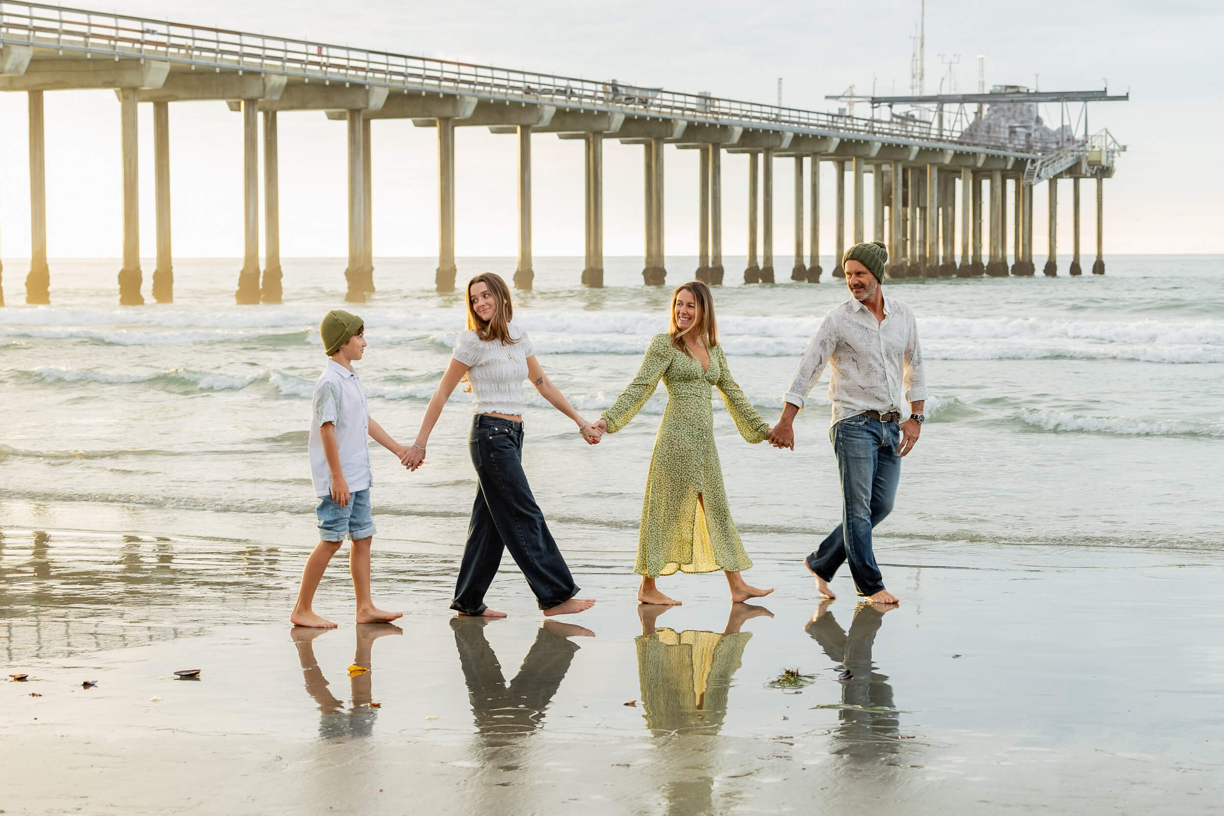 Family holding hands and walking on the beach near the ocean with a pier in the background during sunset.