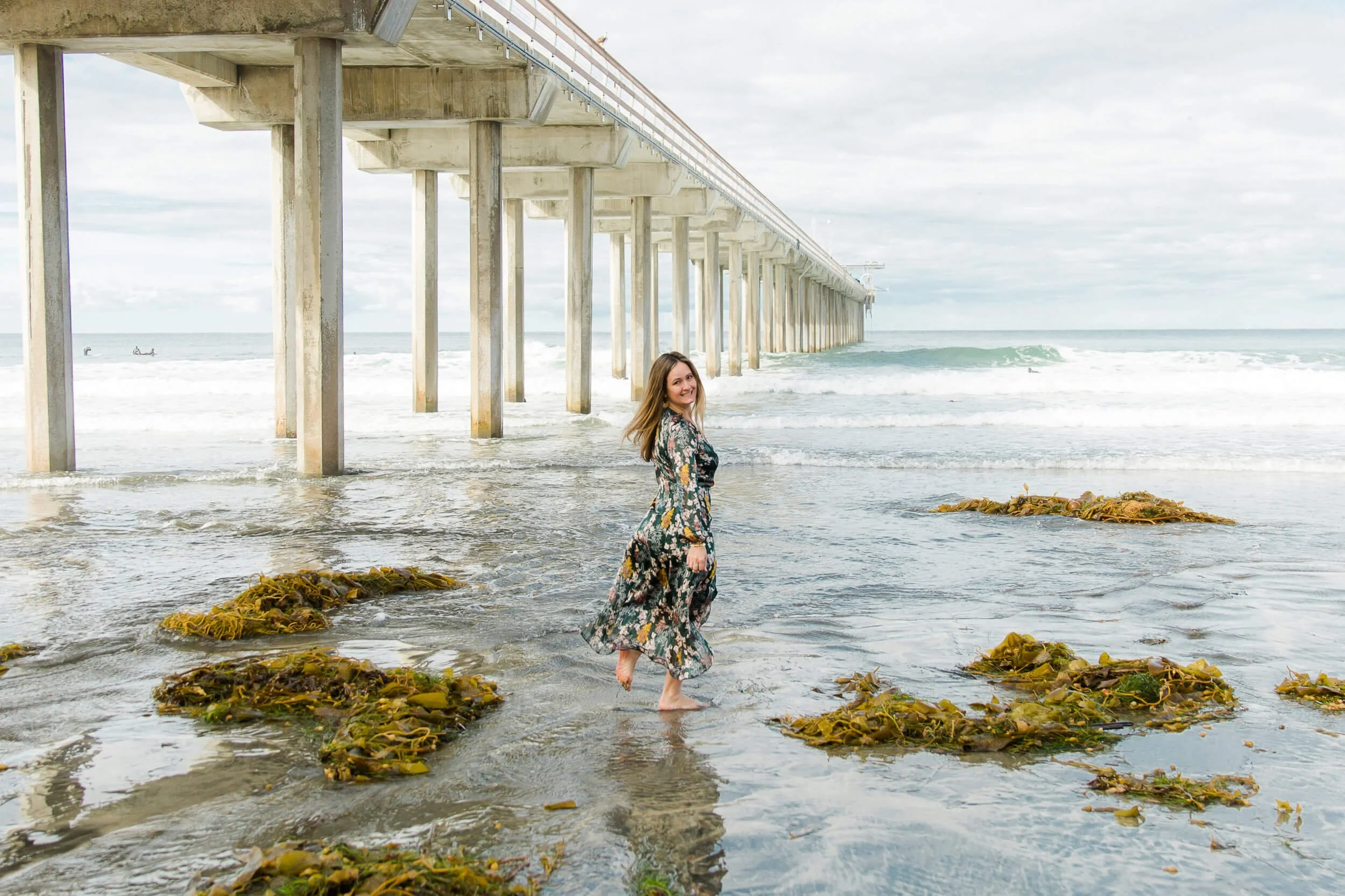 A woman in a floral dress walking in the shallow ocean water under a pier with boats and waves in the background.