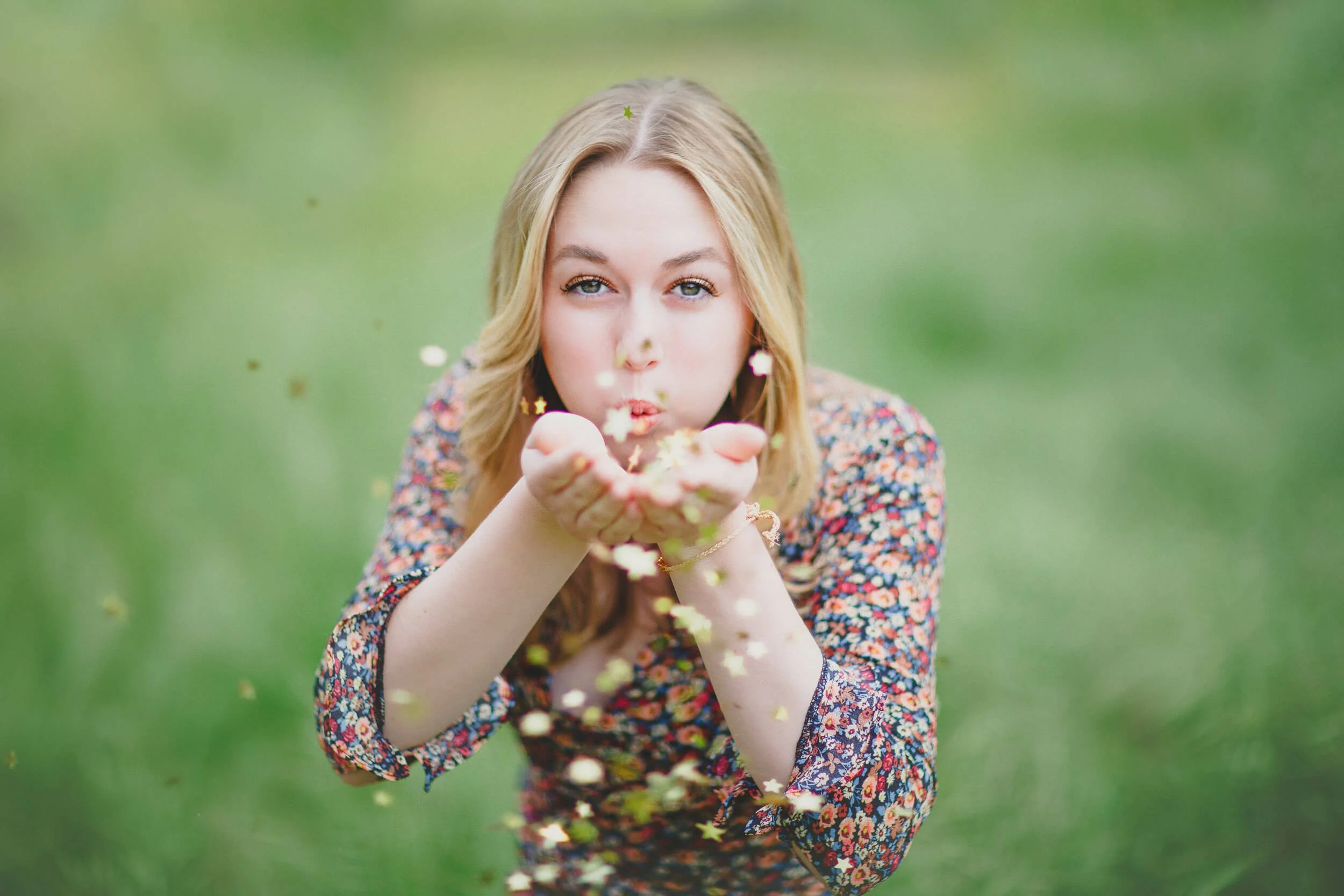 Young woman blowing star-shaped and circular confetti outdoors on a grassy background.
