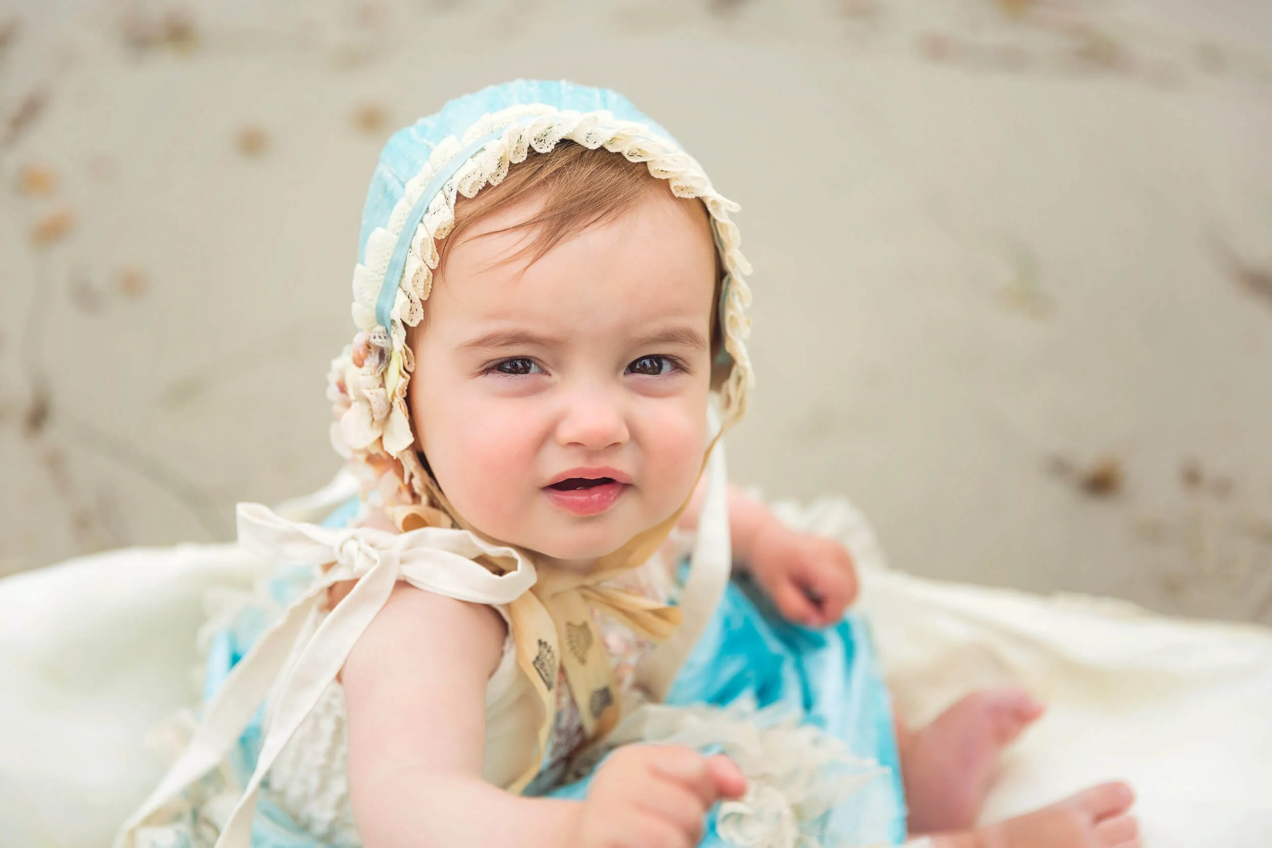 Close-up of a young child with brown hair, wearing a bonnet and summer clothes, sitting on the beach with sand in the background, looking at the camera.