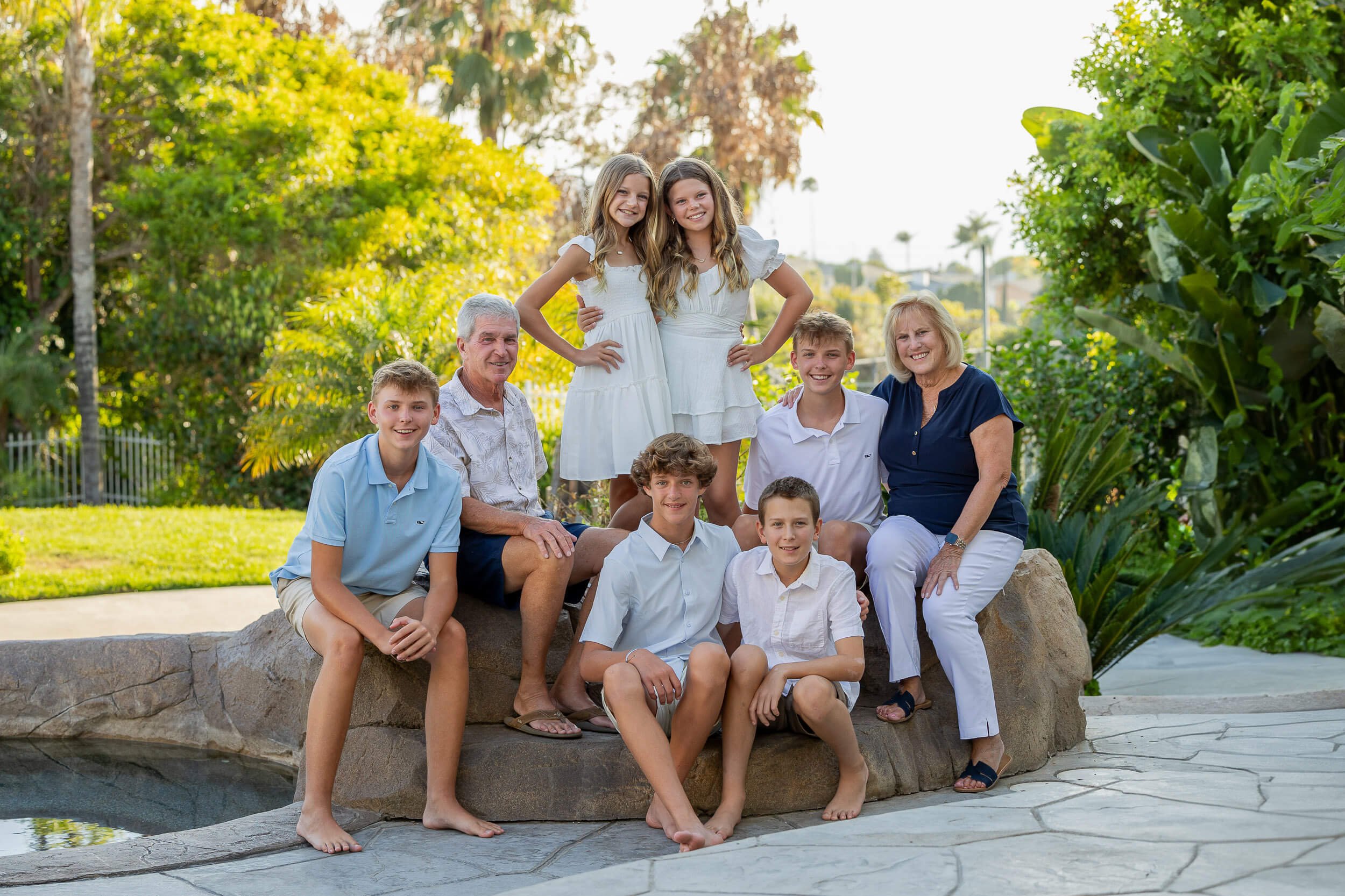 A multi-generational family portrait of nine members outdoors in a lush garden. The group includes three elderly adults, two young adult women, and four children, all smiling and dressed in casual summer clothing. They are gathered around a large roc