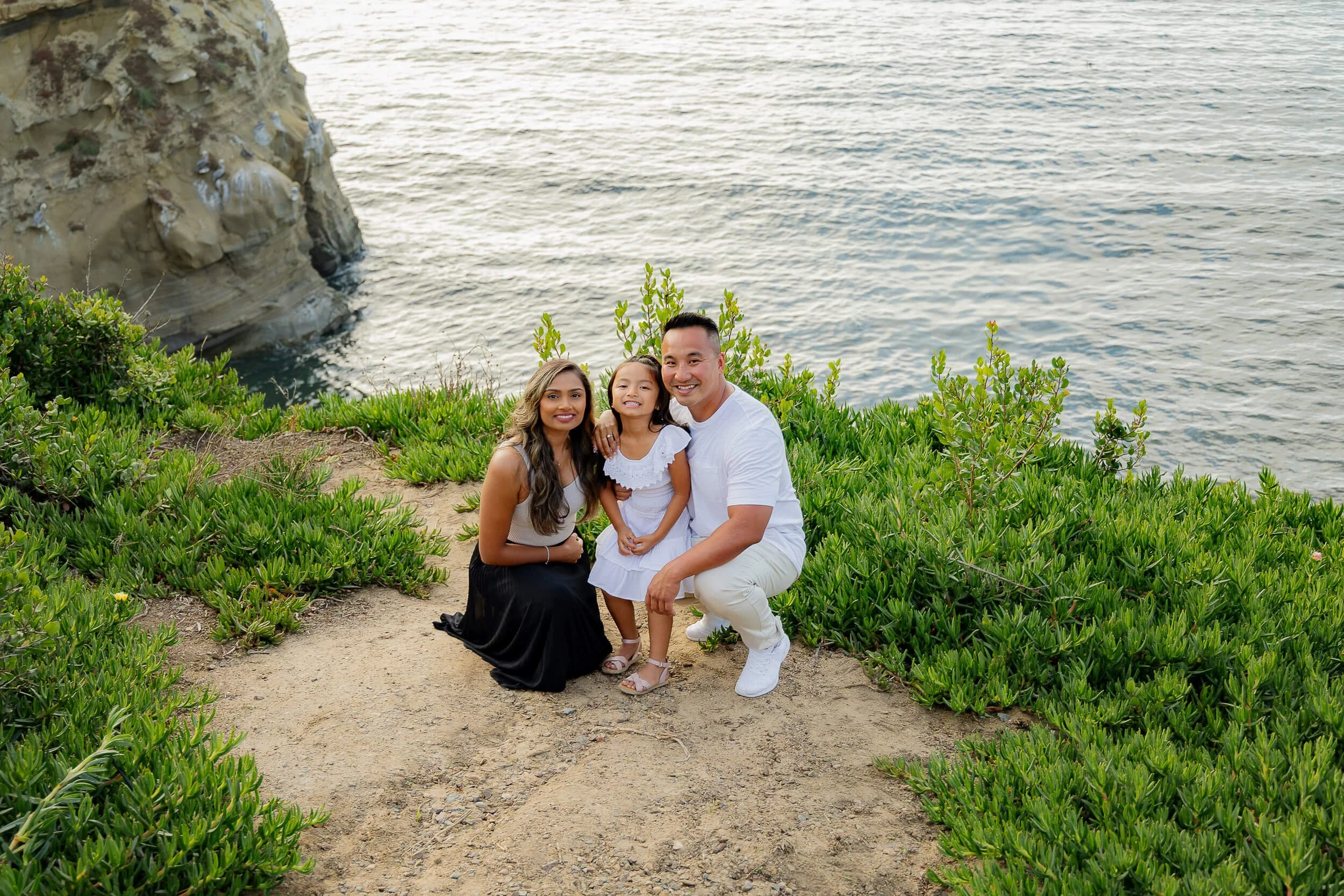 A family of three, a woman, a man, and a young girl, sitting on a dirt path near the ocean with lush green bushes. The woman has long dark hair, and the man and girl have dark hair. All three are smiling at the camera.