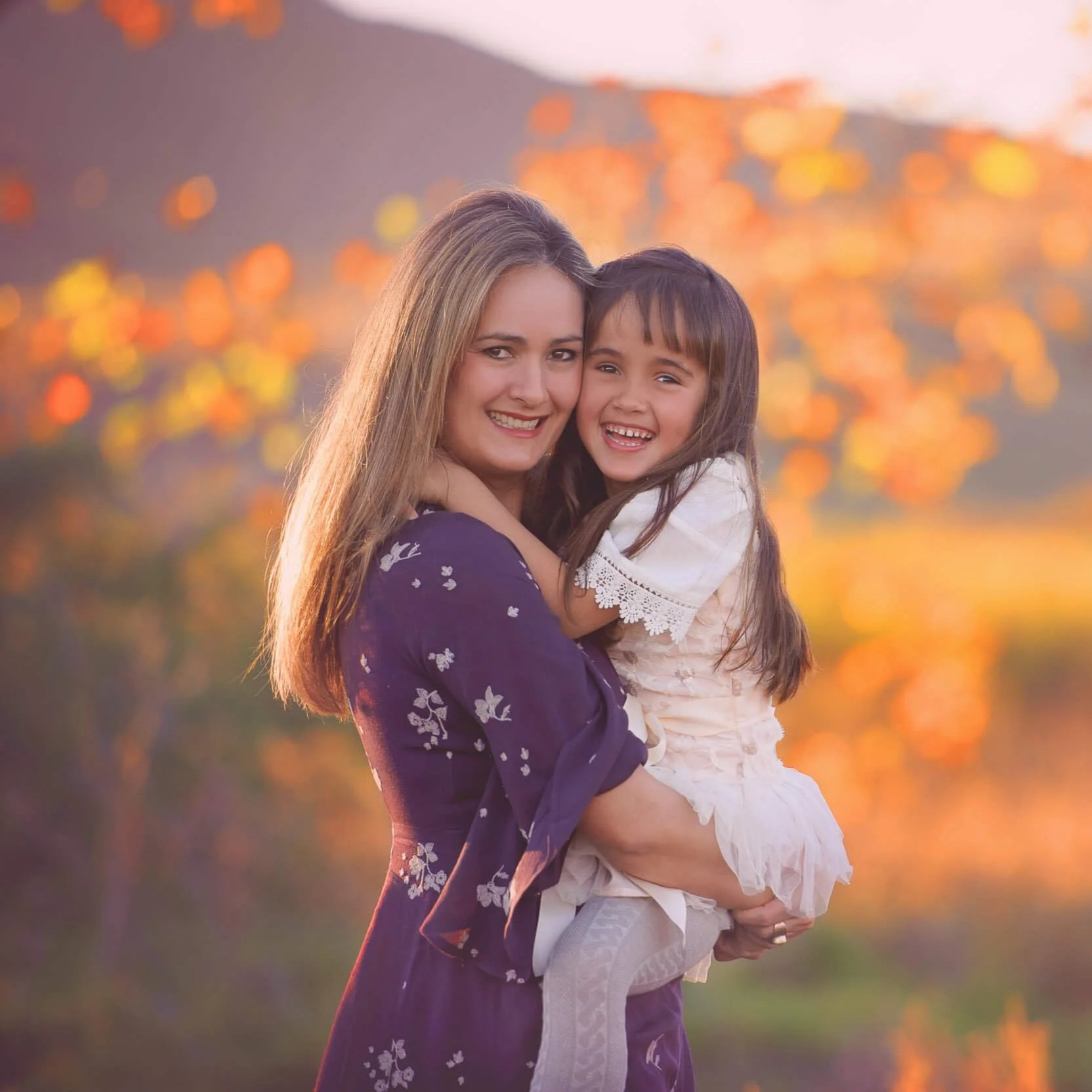 A woman holding a young girl in her arms outdoors during sunset, with colorful fall foliage in the background.