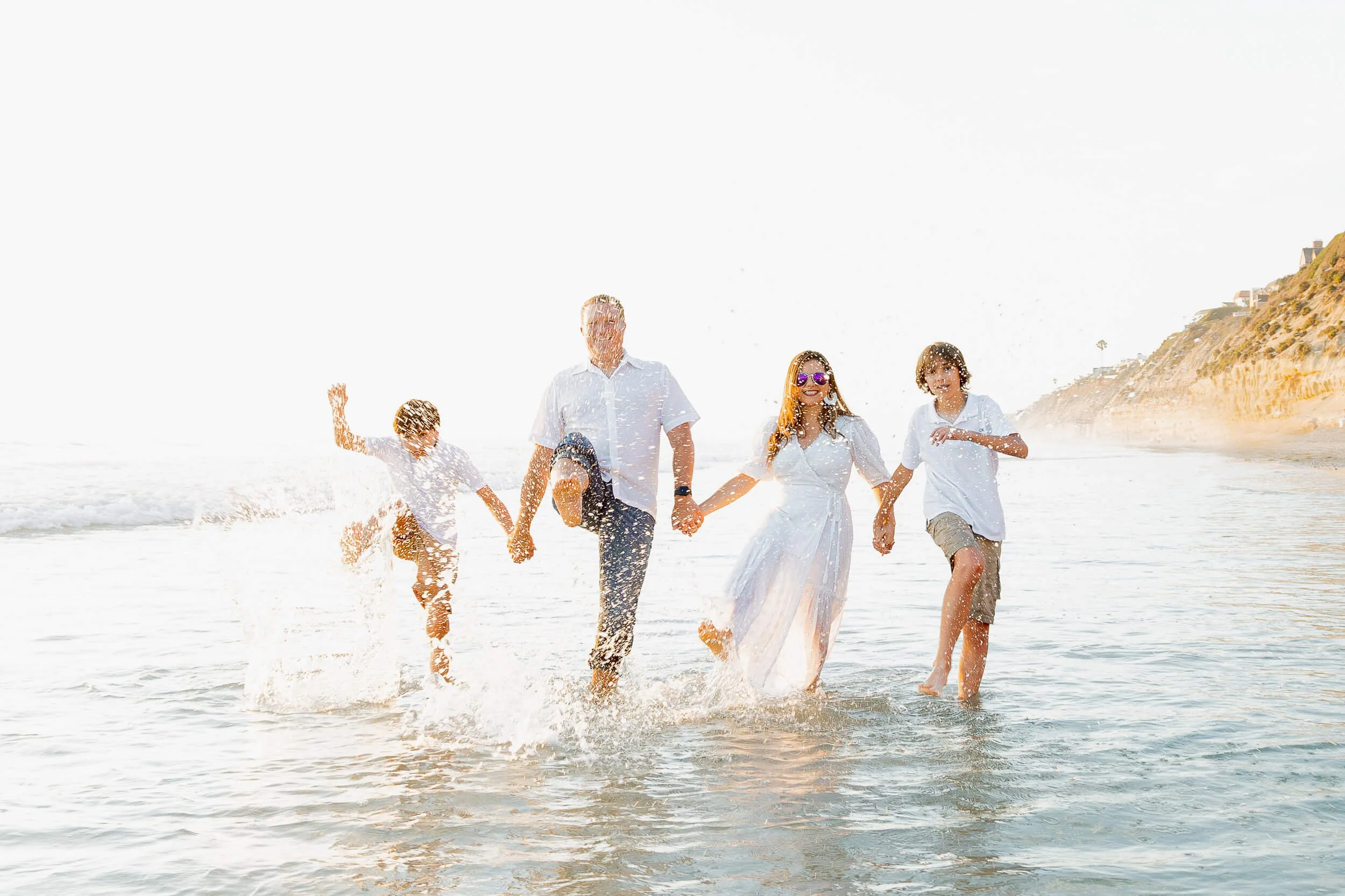 A family of five running through the shallow beach water, holding hands, smiling, with a bright sky and cliffs in the background.