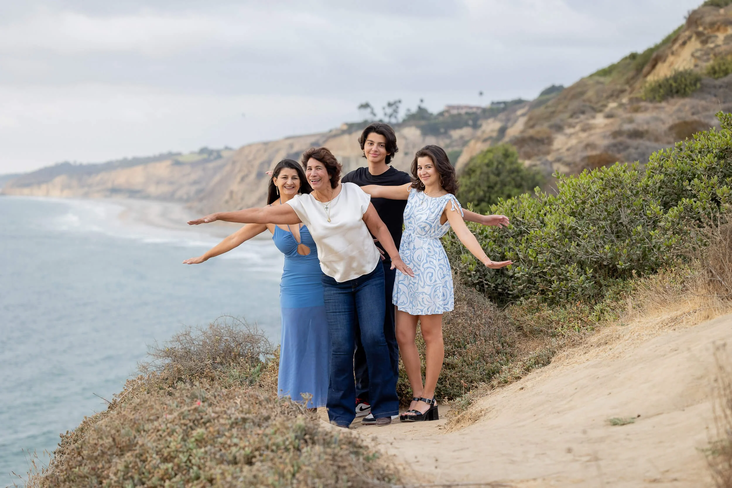 Four women standing on a cliff overlooking the ocean, posing with arms extended and smiling.
