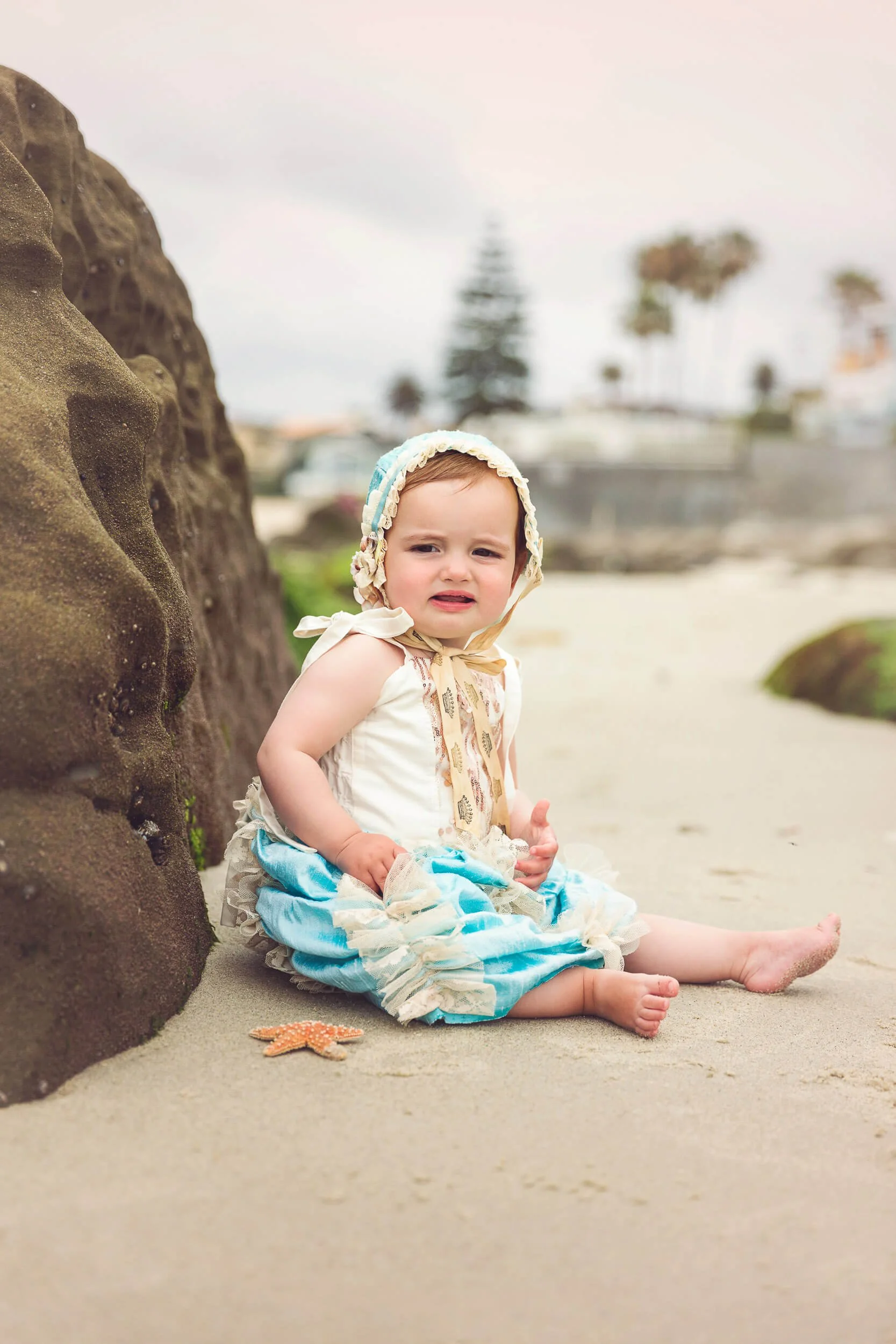 A young girl sitting by a large rock on a sandy beach, wearing vintage-style clothing and a bonnet, with a small starfish nearby, and a blurred background of palm trees and overcast sky.