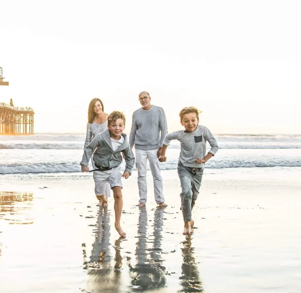 A family of five enjoying a day at the beach, running in the shallow surf with a pier in the background during sunset.