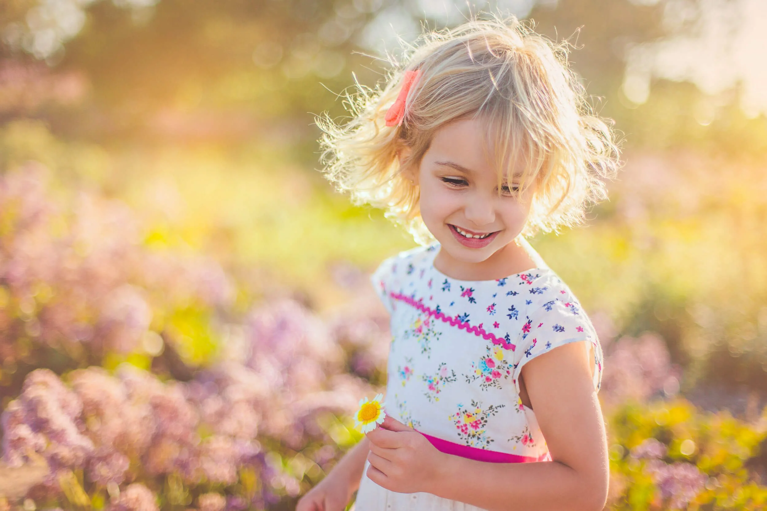 A young girl with blonde wavy hair, wearing a white dress with colorful floral patterns, holding a small yellow daisy flower, standing outdoors in a field of purple and pink flowers with sunlight in the background.