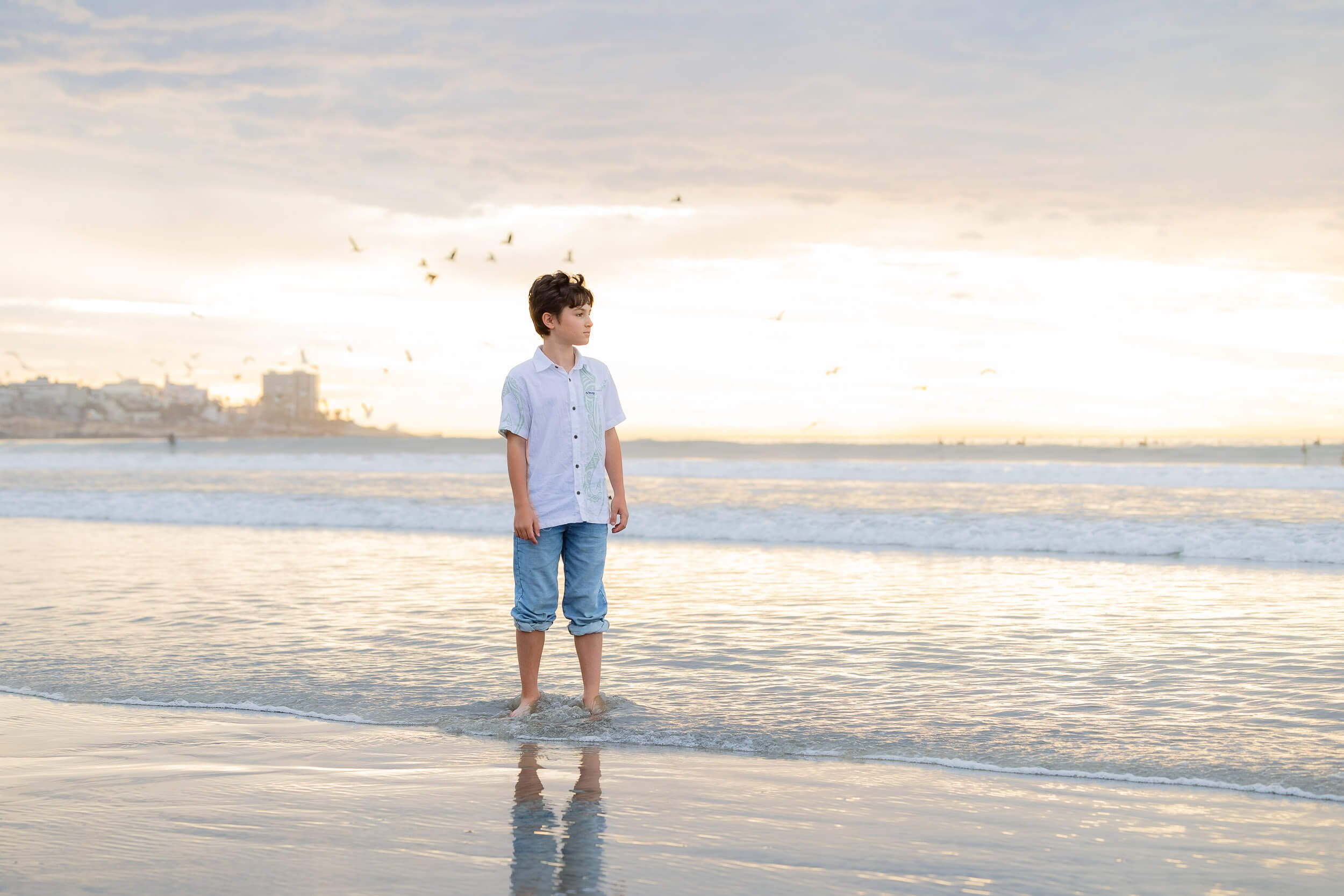 A boy with dark hair wearing a white shirt and rolled-up jeans standing in shallow ocean water at sunset, with birds flying in the sky in the background.