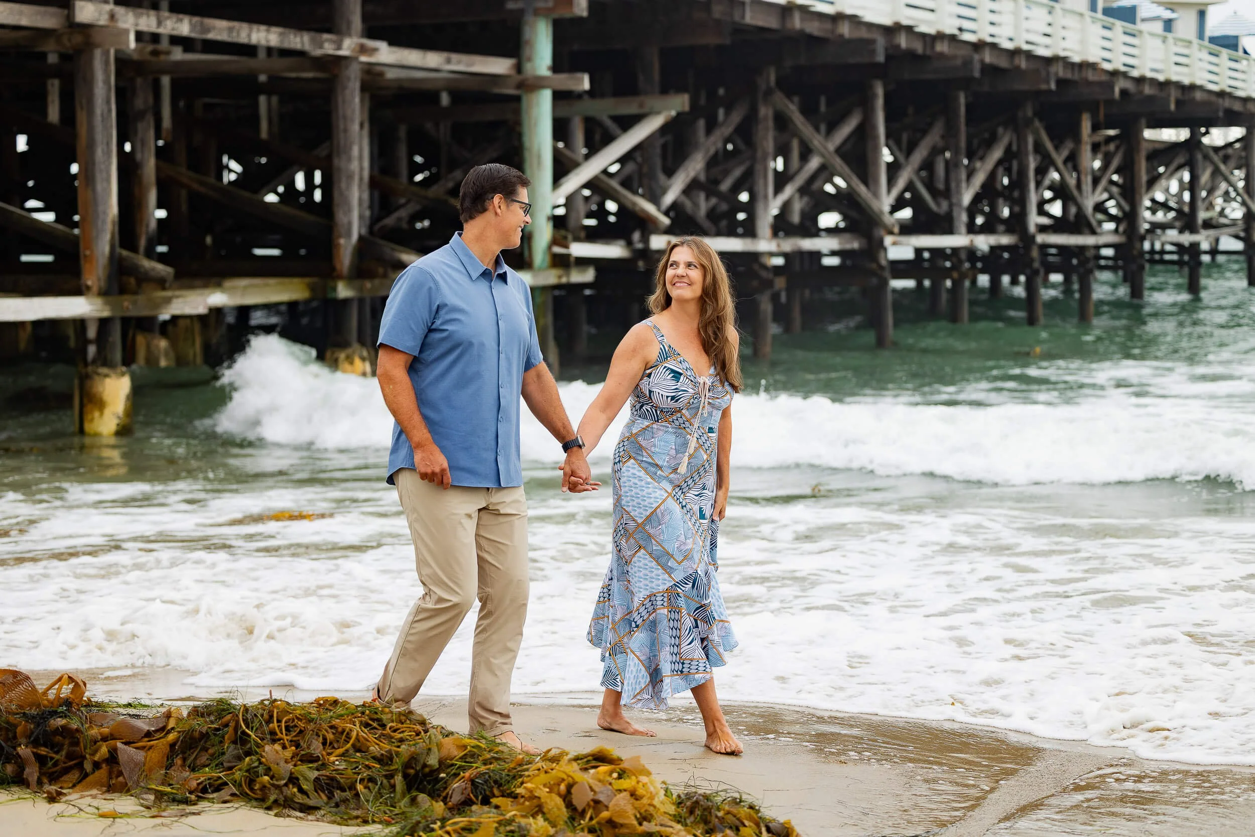 A couple walking hand in hand along the beach, holding hands and smiling, near a wooden pier with waves crashing on the sandy shore.