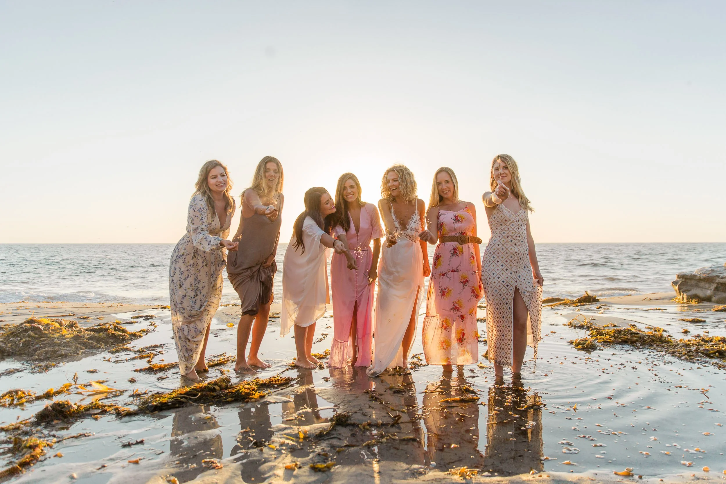 Group of seven women in dresses standing by the ocean at sunset, holding sparklers and smiling.