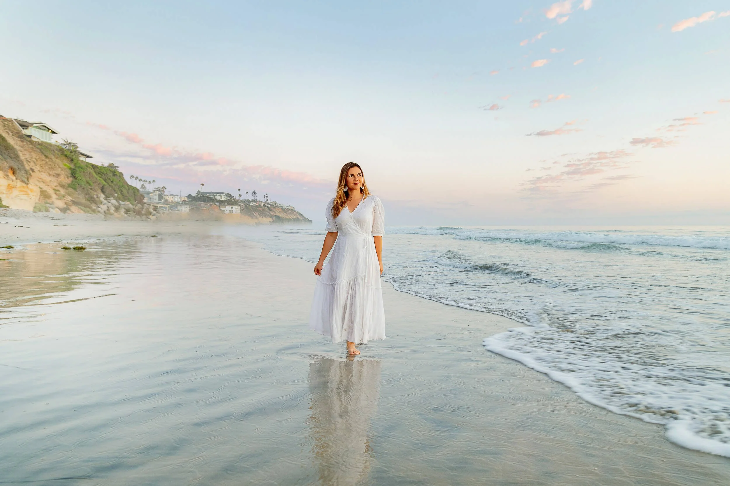 Woman in a white dress walking along the beach at sunset.