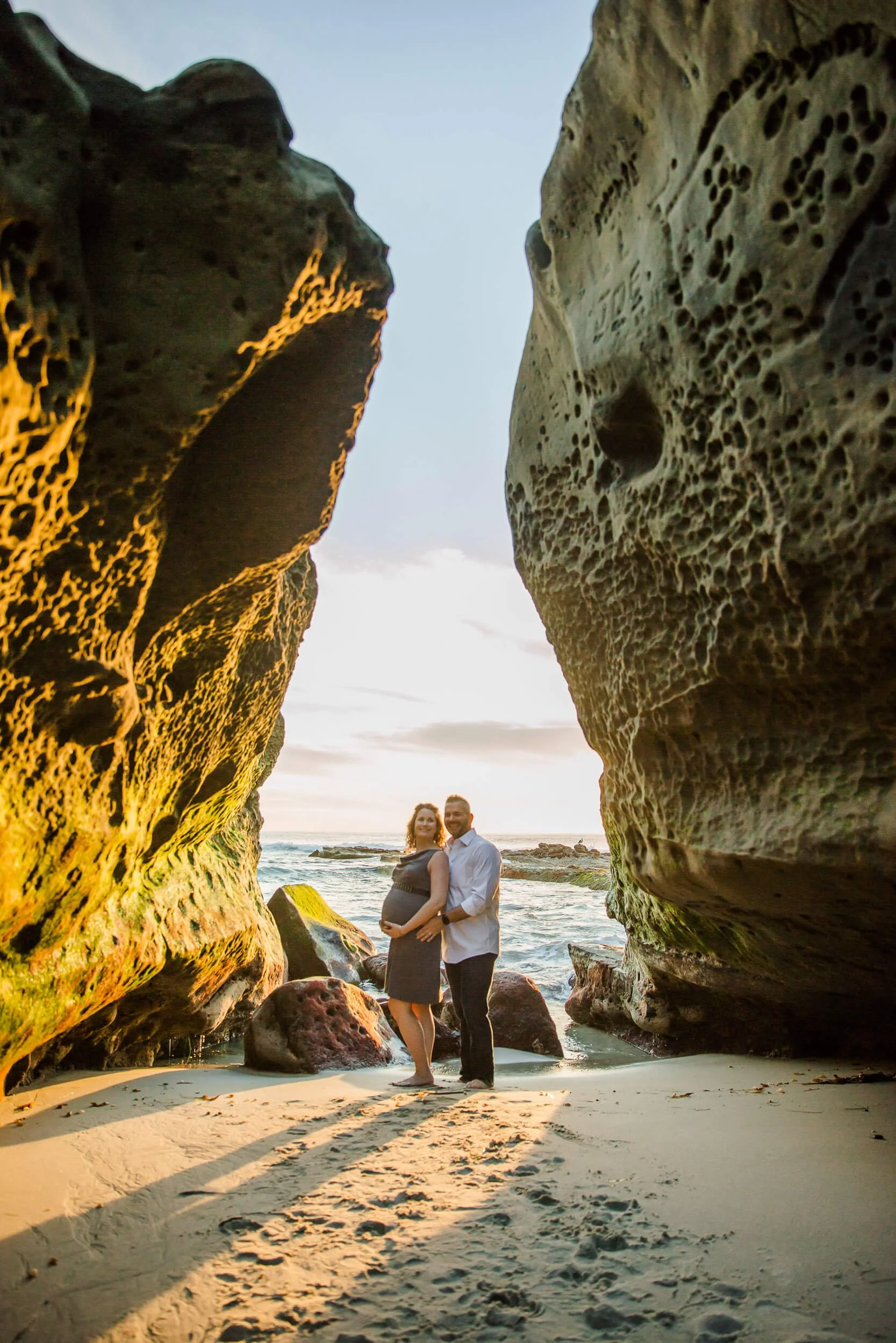 A couple standing between large rocky formations on a sandy beach at sunset, with the ocean in the background.