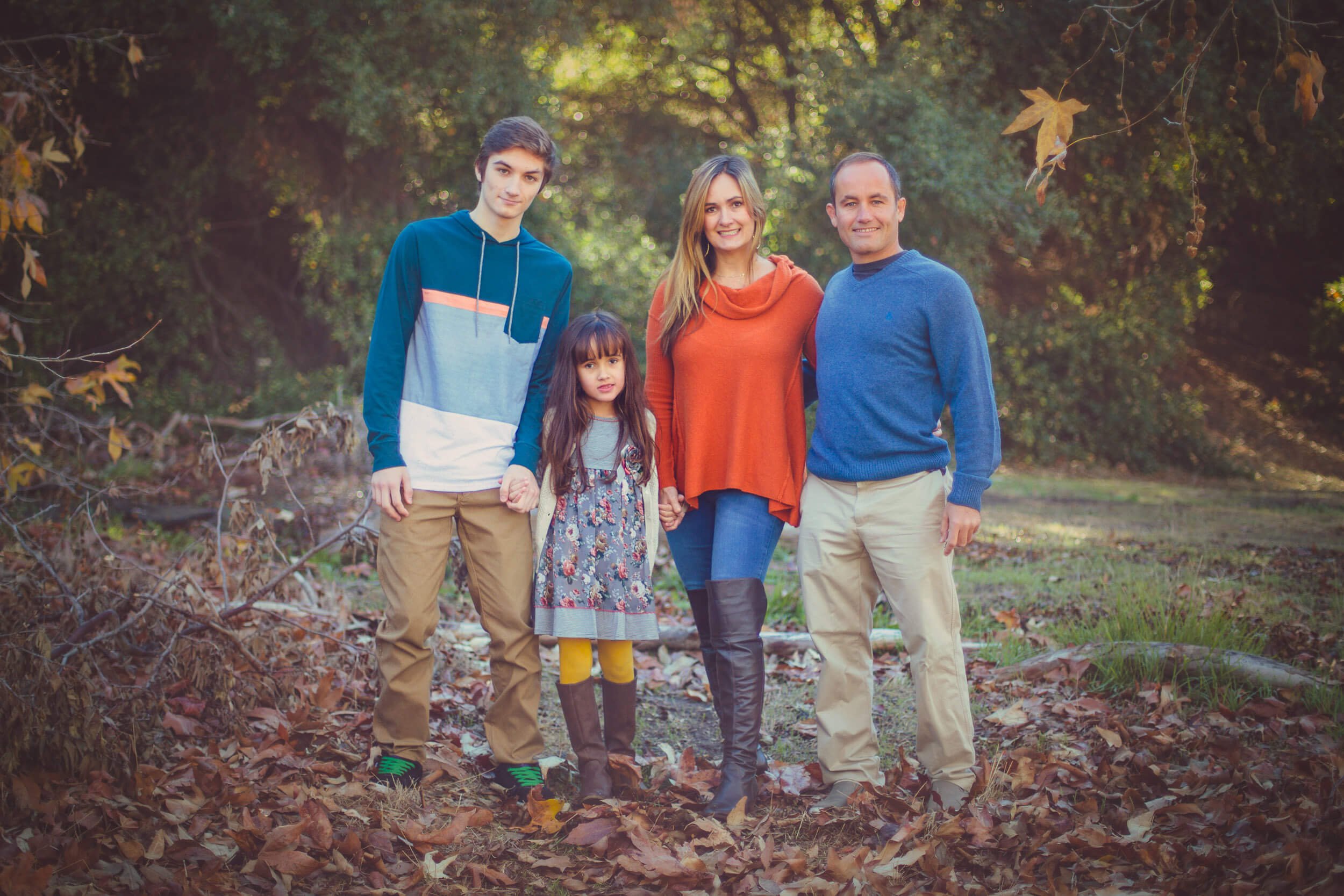 A family of five standing outdoors on a fall day, surrounded by leaves and trees, holding hands and smiling.