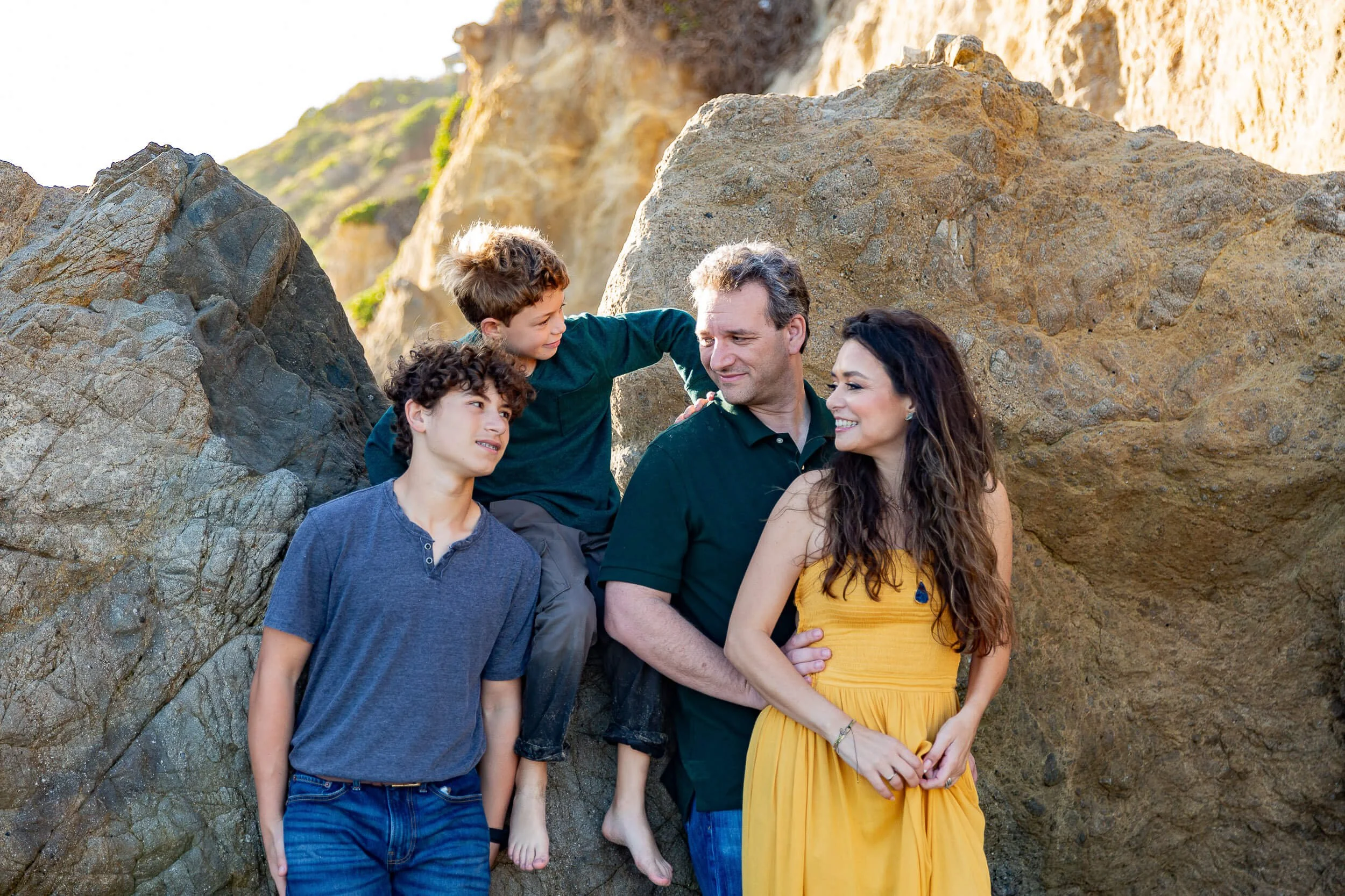 A family of five standing on rocks outdoors during daytime, smiling and looking at each other, with a sunlit rocky landscape in the background.