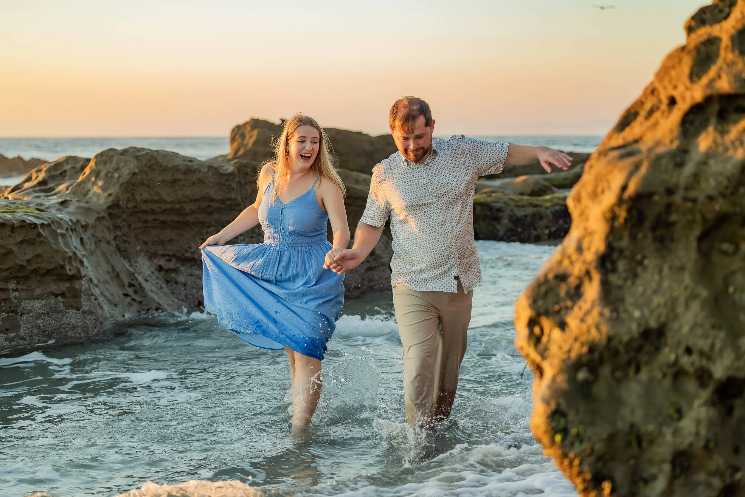 A young woman in a blue dress and a young man in a patterned shirt and beige pants are happily holding hands and playing in the shallow ocean water near rocks at sunset.