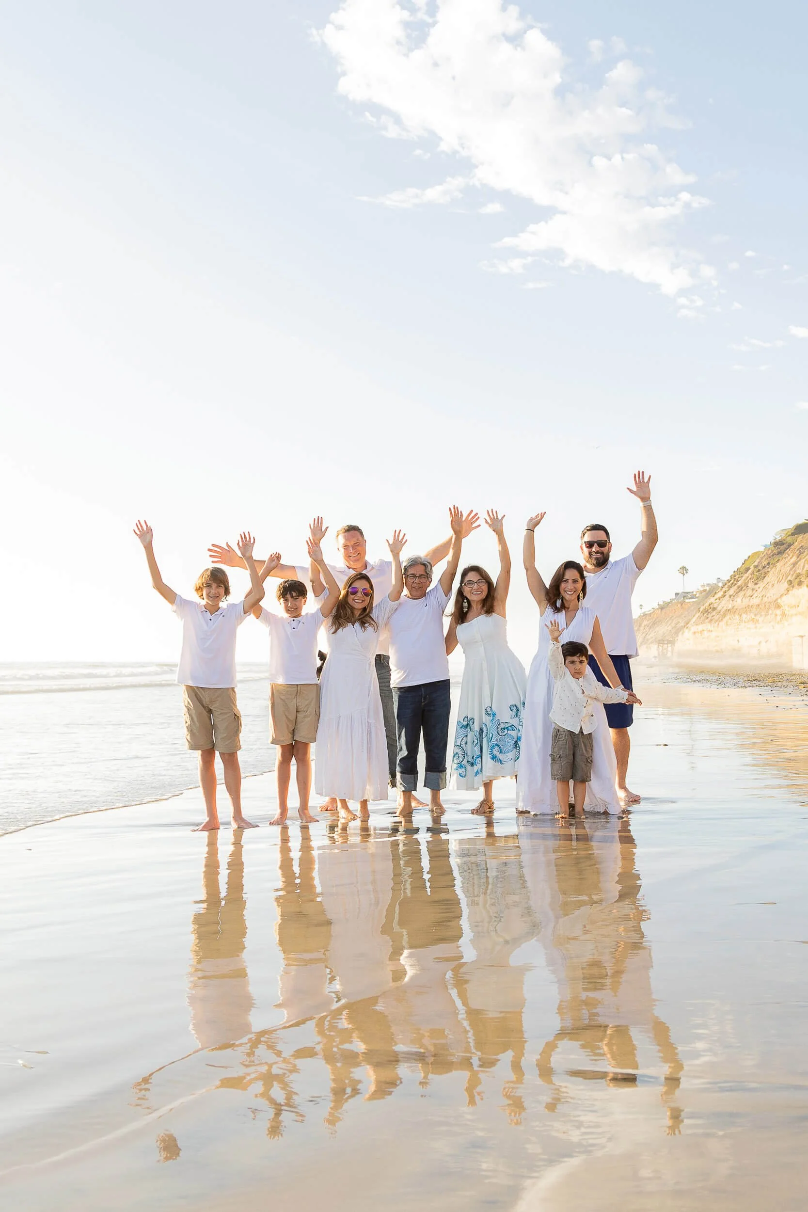 Family of nine enjoying a walk on the beach with their hands raised, smiling, under a partly cloudy sky during sunset.