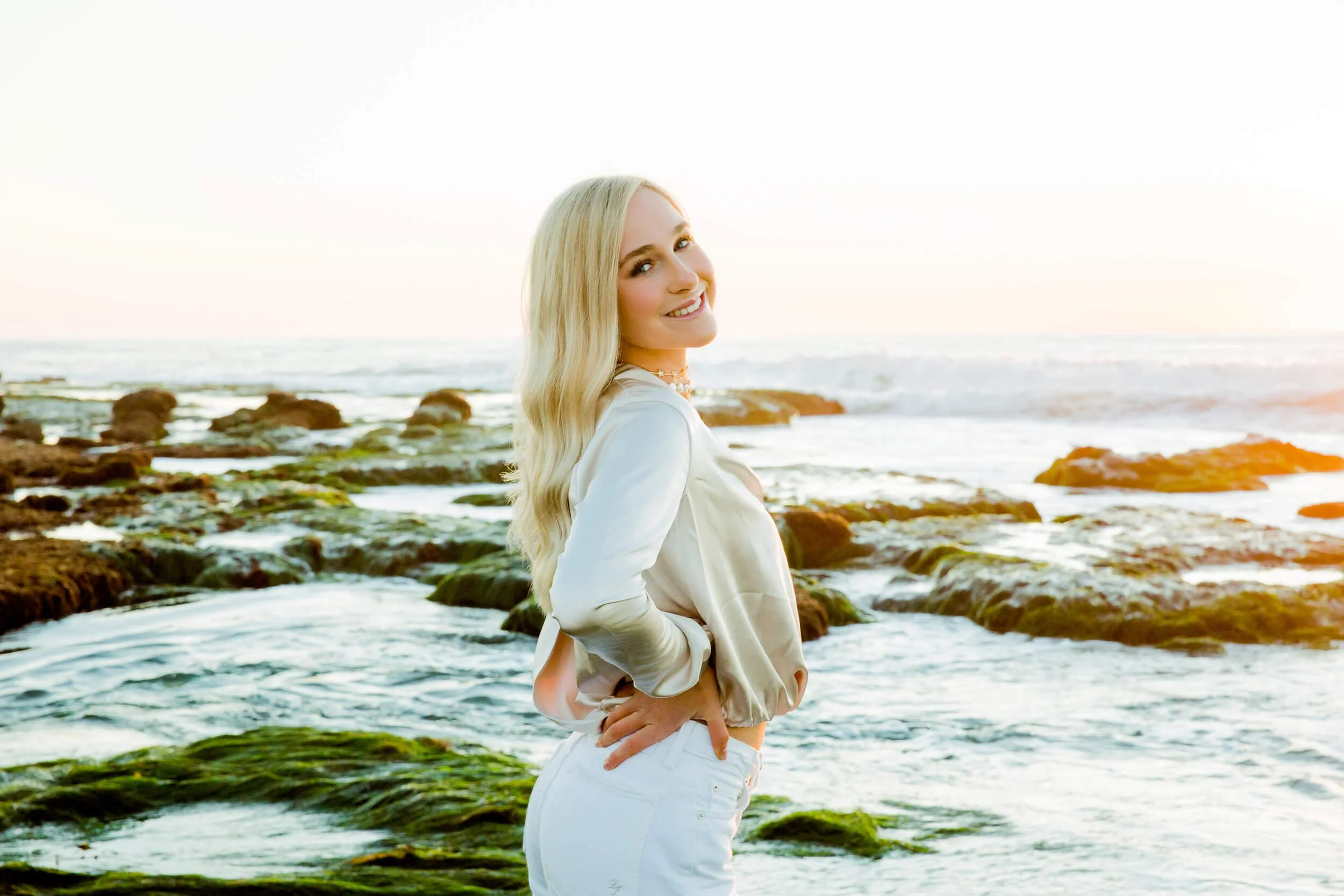 A woman with long blonde hair standing on a rocky beach at sunset, smiling and looking at the camera.