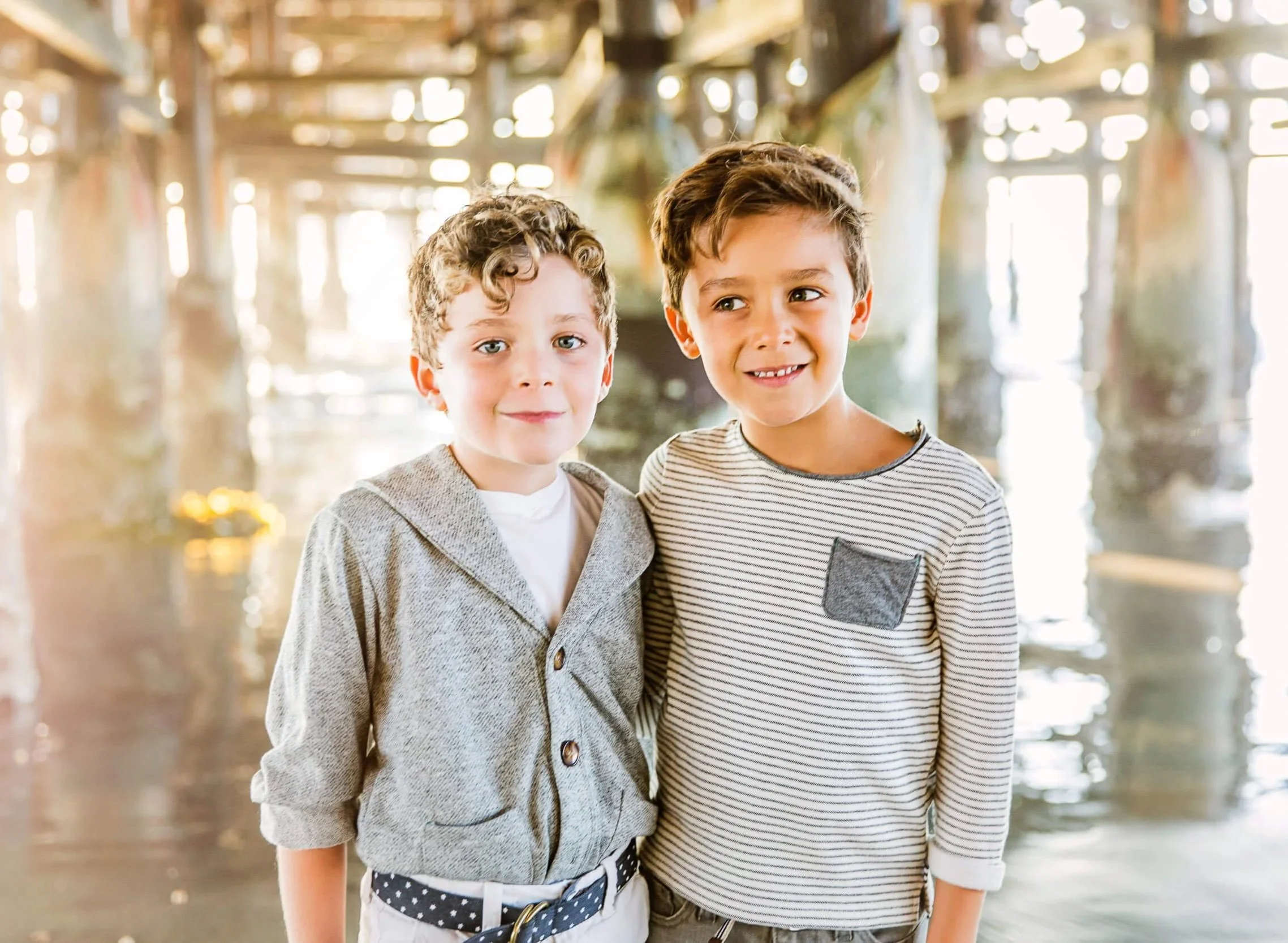 Two young boys standing close together under a pier or bridge over water, smiling and looking at the camera.