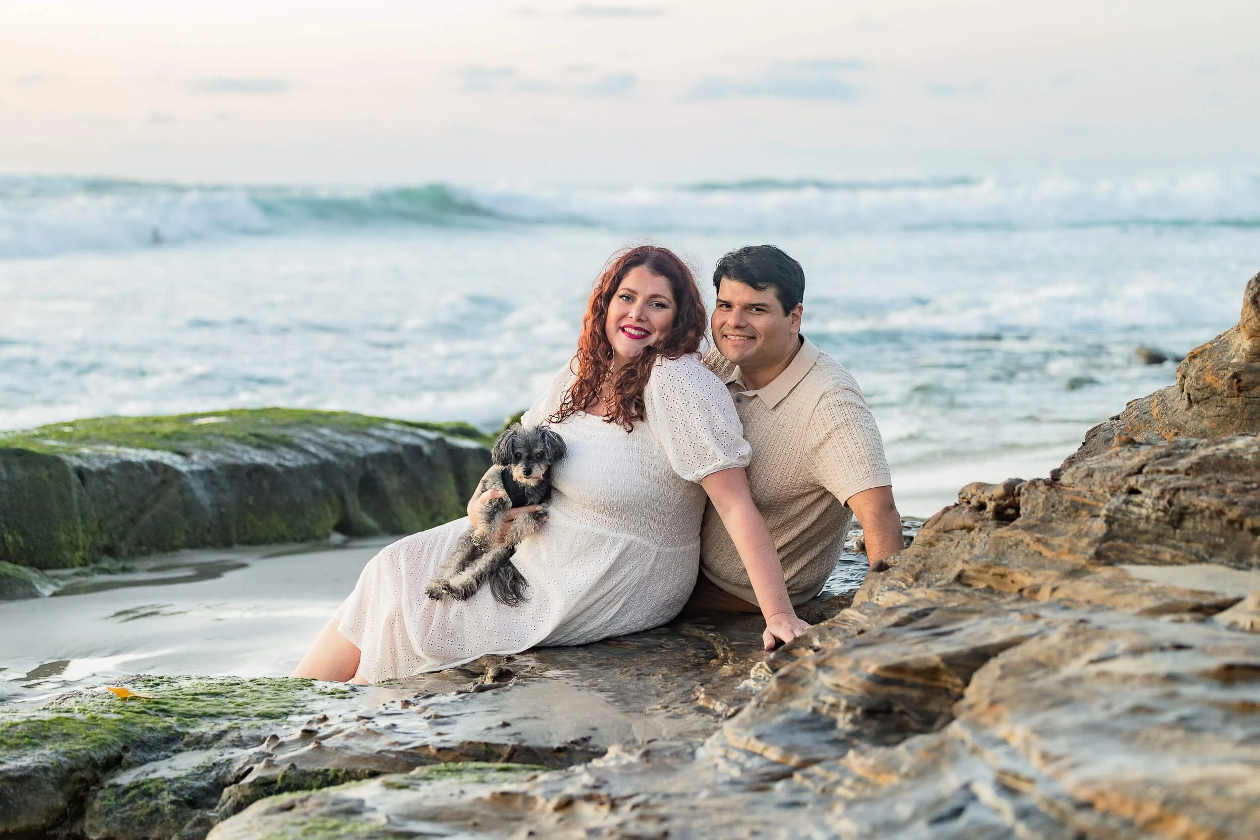 A smiling couple sitting on rocks at the beach with their small black and gray dog, ocean waves in the background, during daylight.