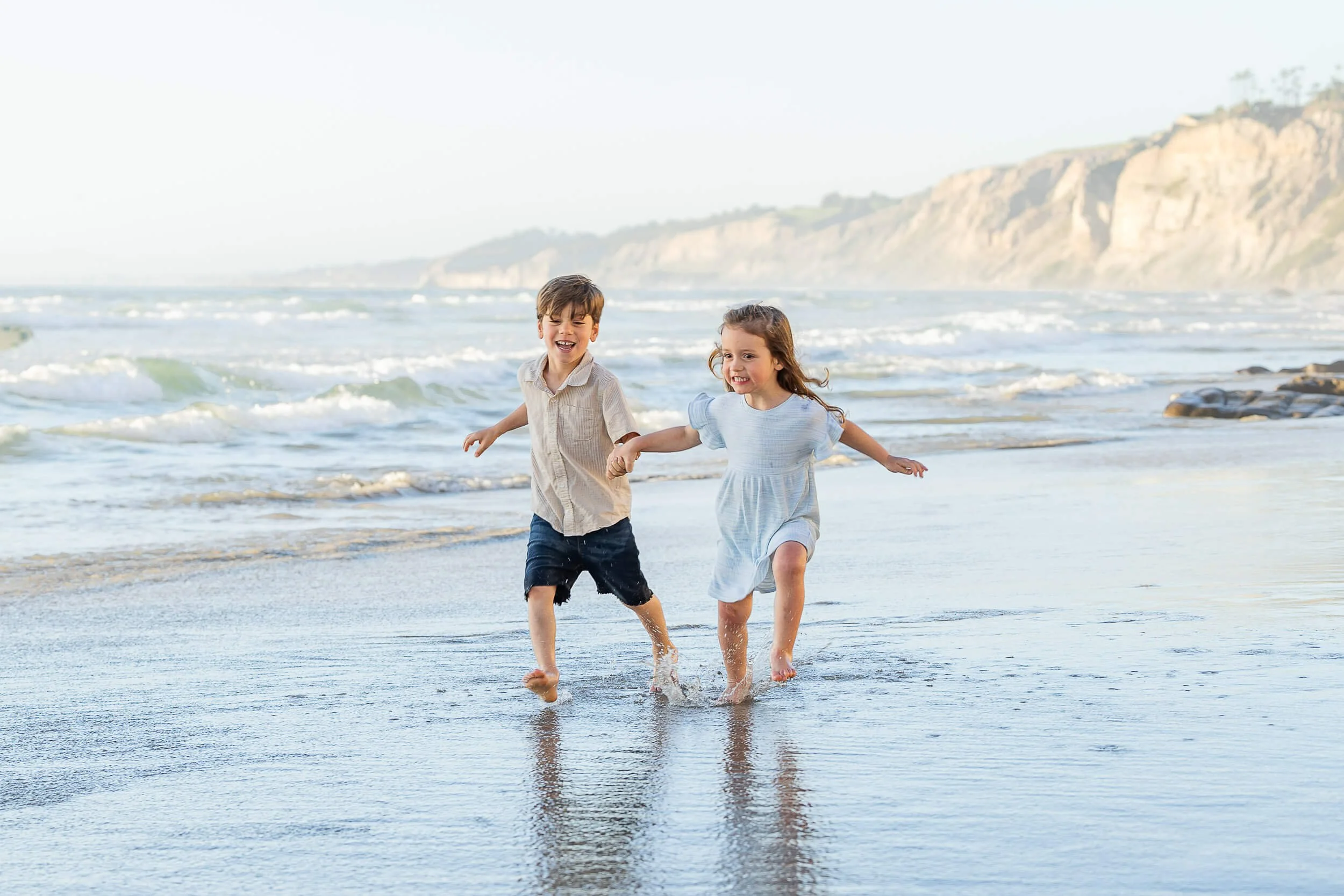 Two children, a boy and a girl, playing and running barefoot along the shoreline at the beach, holding hands and smiling. Cliffs in the background, ocean waves, clear sky.