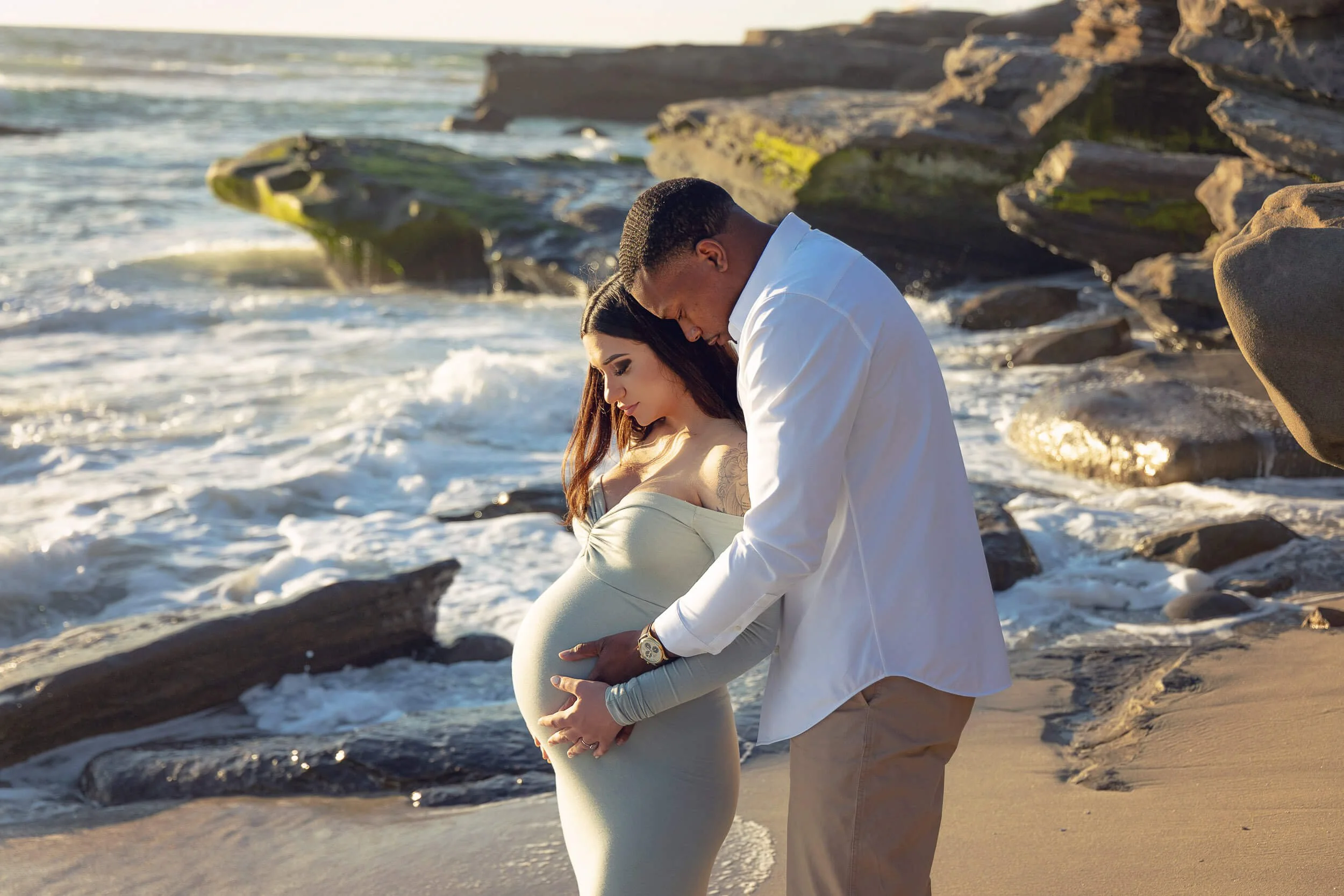 A pregnant woman and her partner standing on a beach, holding her belly. The man is touching her stomach with both hands while the woman looks down at her belly. Waves crash on rocks in the background during sunset.