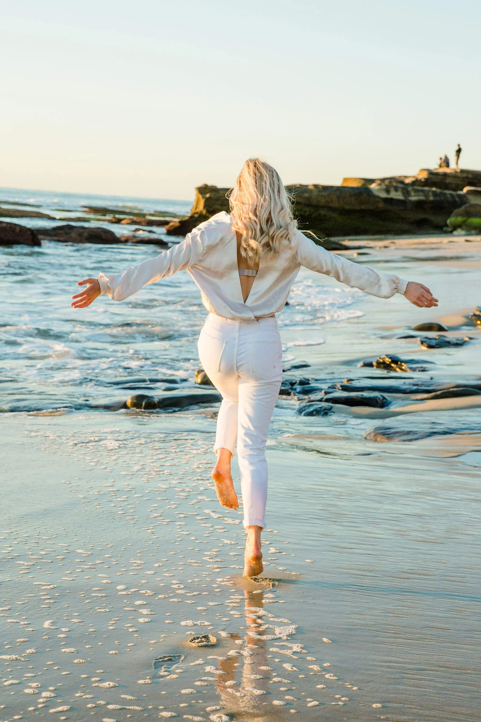 Woman in white clothing running along the beach with arms outstretched during sunset.