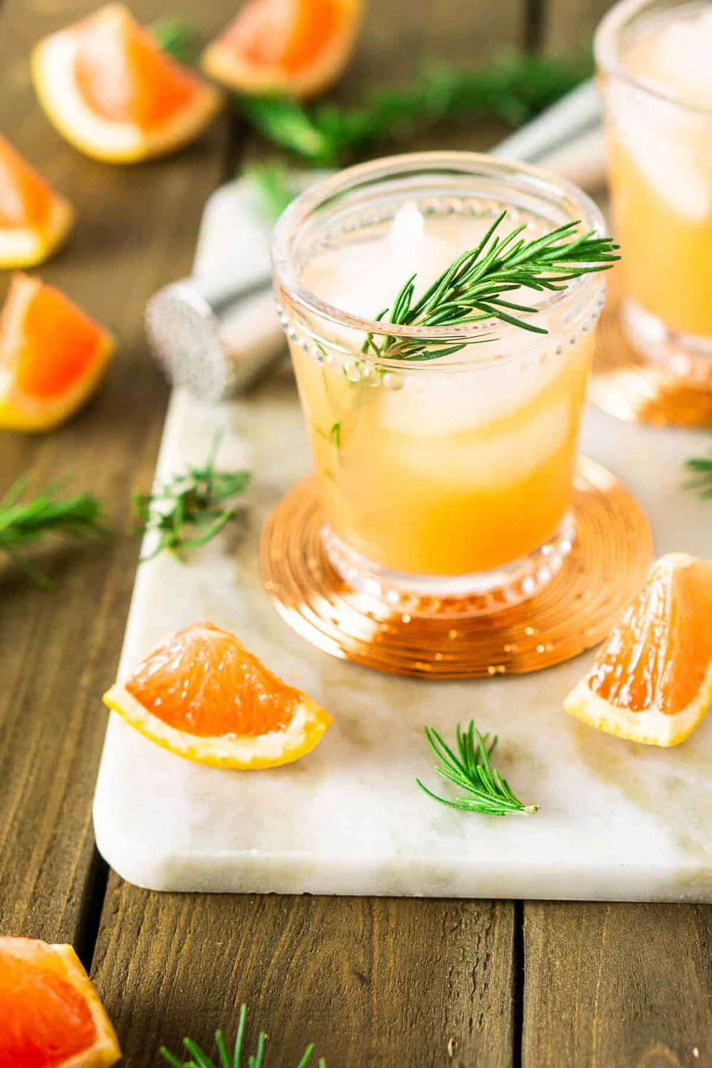 A glass of grapefruit juice garnished with a sprig of rosemary on a white marble board, surrounded by grapefruit slices.