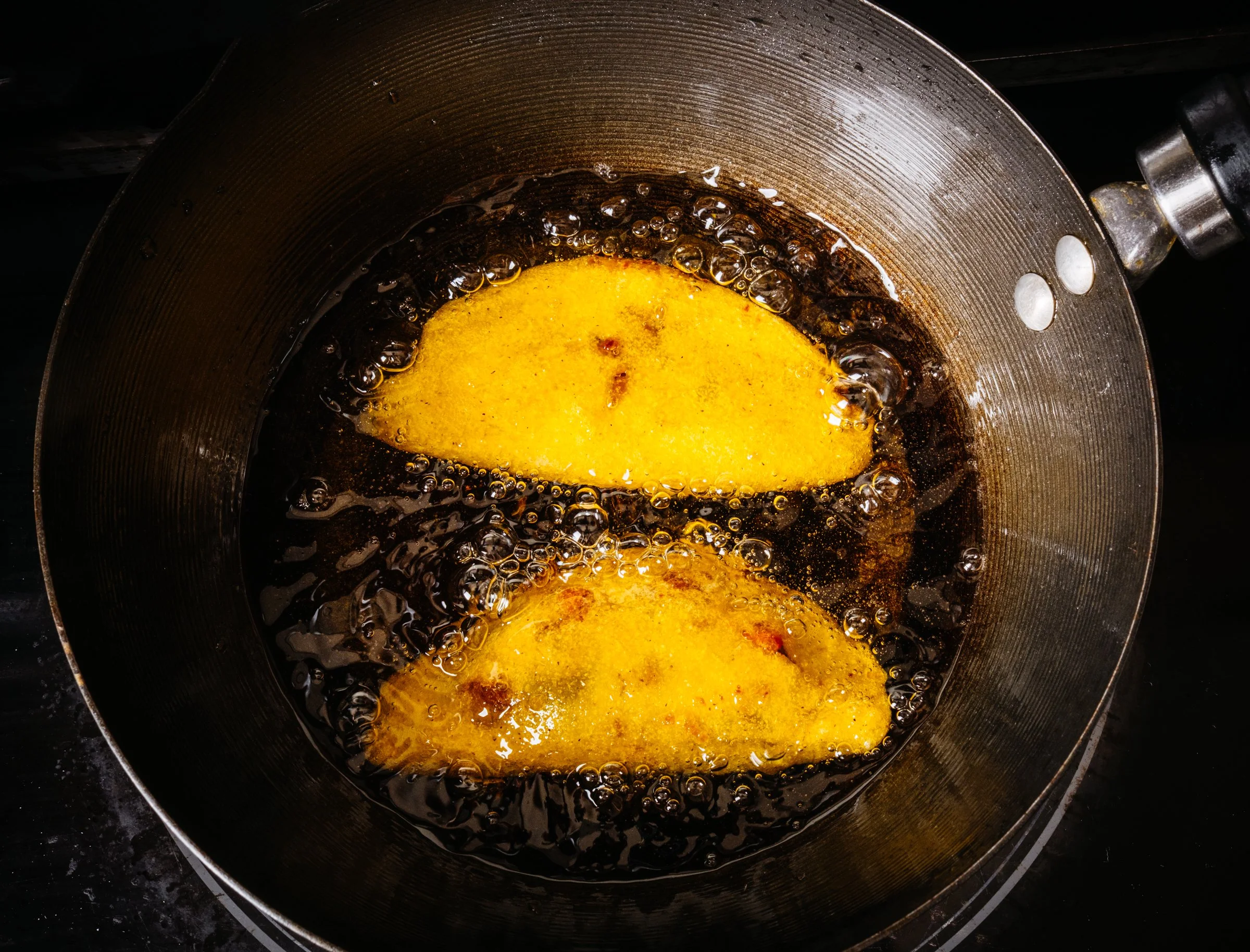 Two pieces of battered and fried plantains frying in a frying pan with bubbling oil.