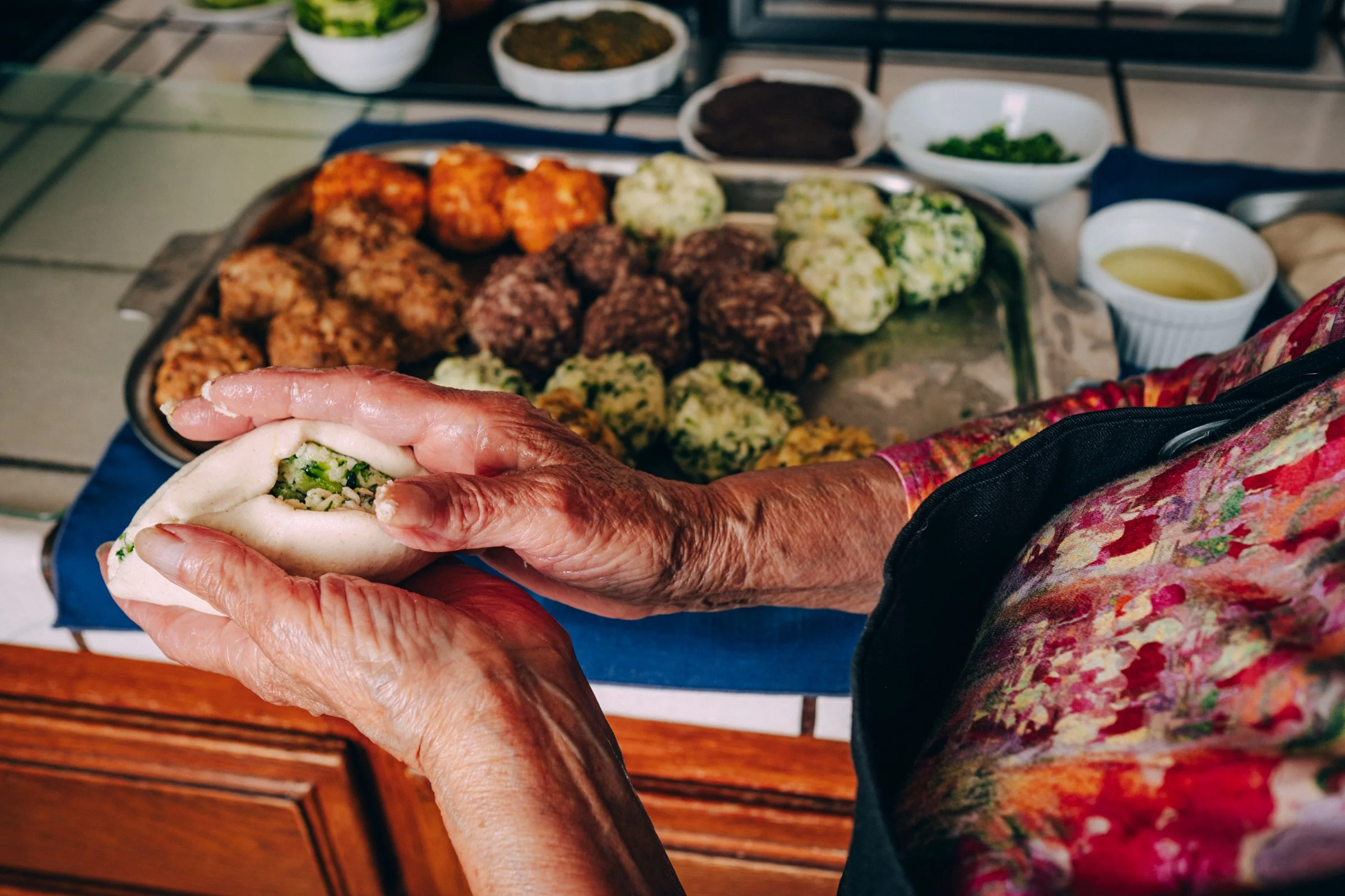 An elderly woman holding a steamed bun filled with chopped vegetables, with a tray of various meat and vegetable dumplings behind her.