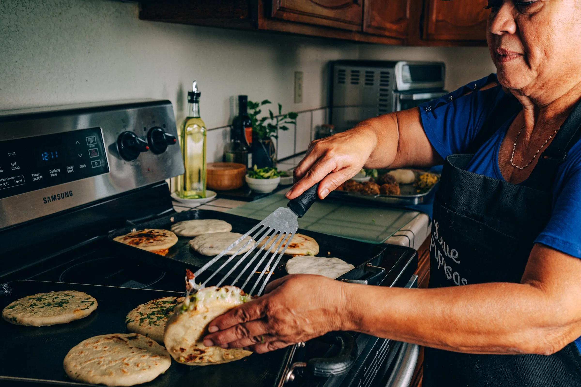 Yanira, the owner and operator of Love Pupusas, a MEHKO in San Diego, cooking on a stove, flipping pupusas with a spatula in a kitchen.