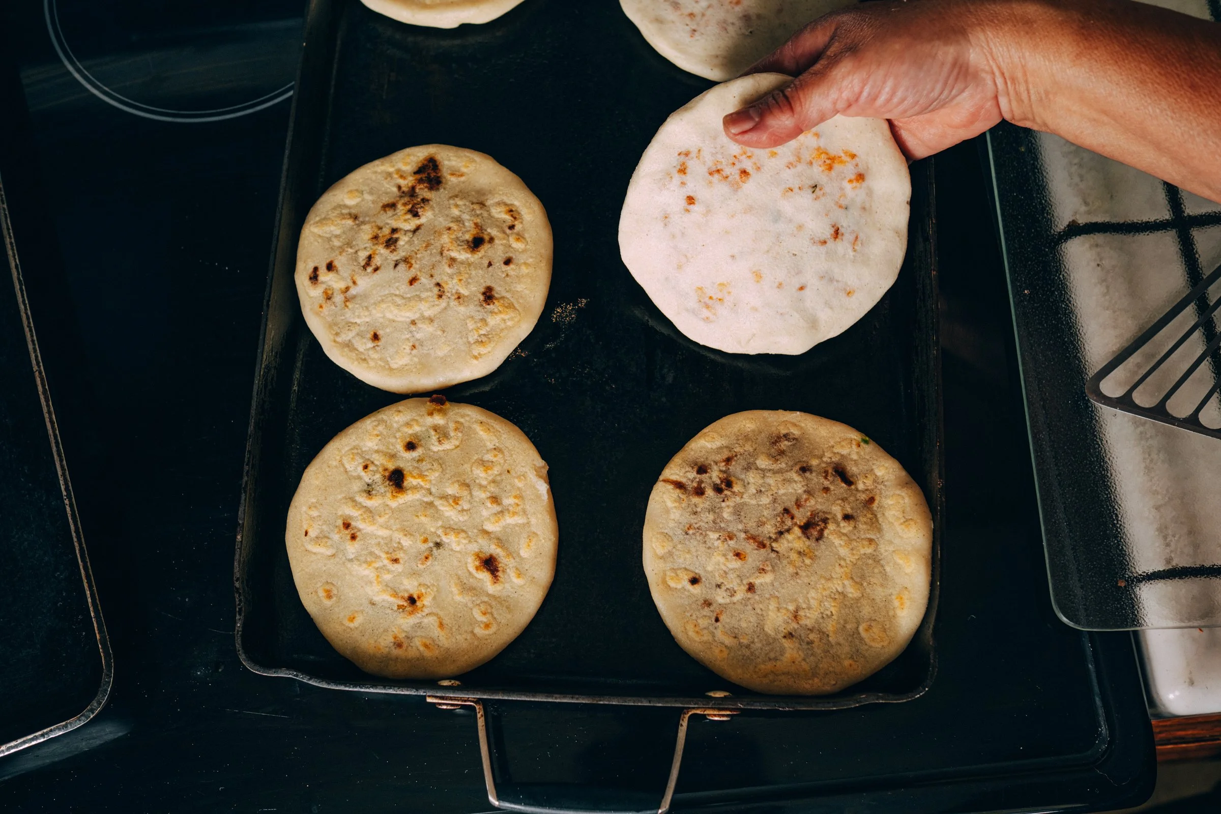 Four pieces of pupusas toasting on a black stovetop, with one being lifted by a hand.