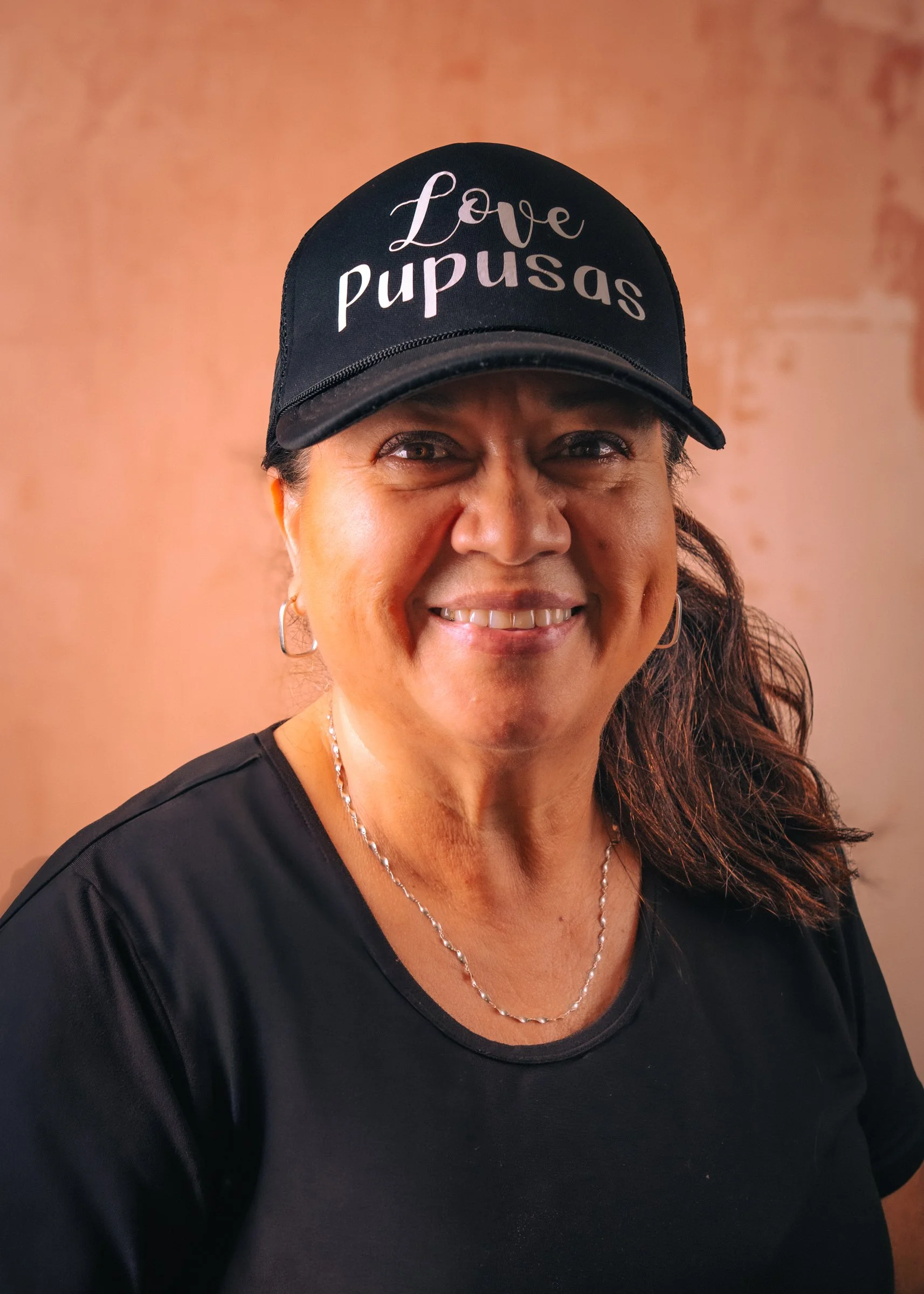 A smiling Yanira, the owner and operator of Love Pupusas, a MEHKO in San Diego wearing a black cap with the words 'Love Pupusas' and a black shirt, standing against a blush background.
