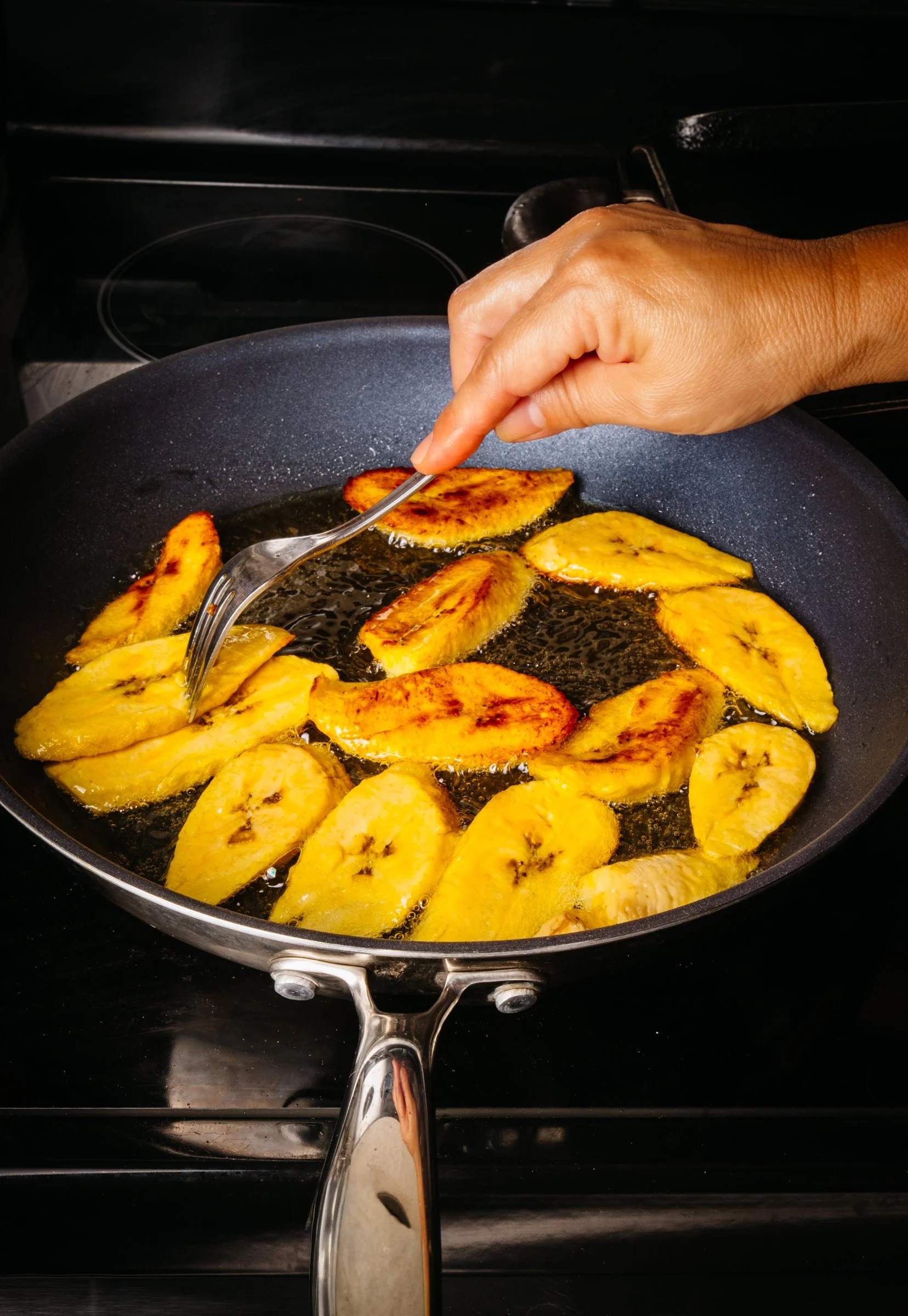 A person is grilling sliced bananas in a black frying pan on a stovetop, using a fork to turn the bananas.
