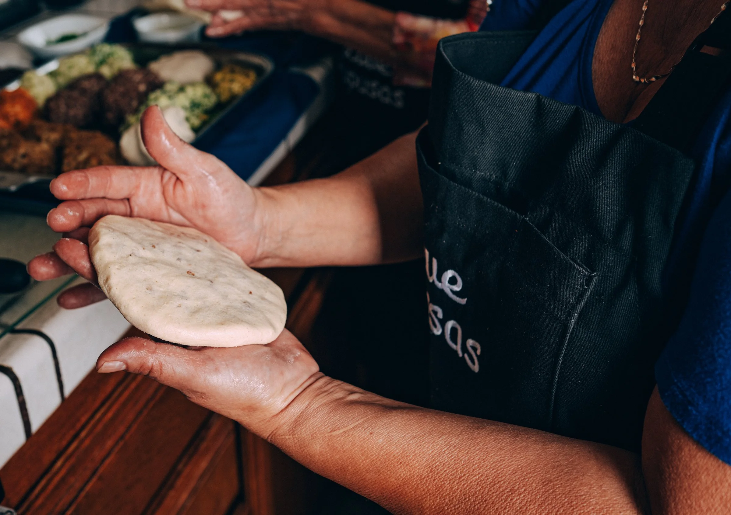 Person holding a piece of flatbread or dough in their hands, with a tray of Food in the background.