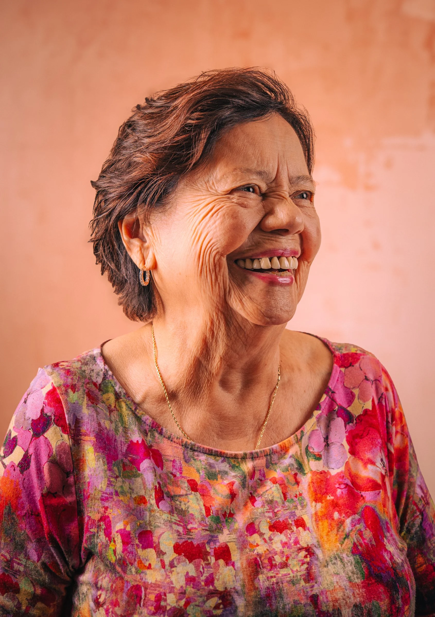A smiling elderly woman with short, dark hair, wearing floral clothing, earrings, and a gold necklace, standing against a soft pink background.