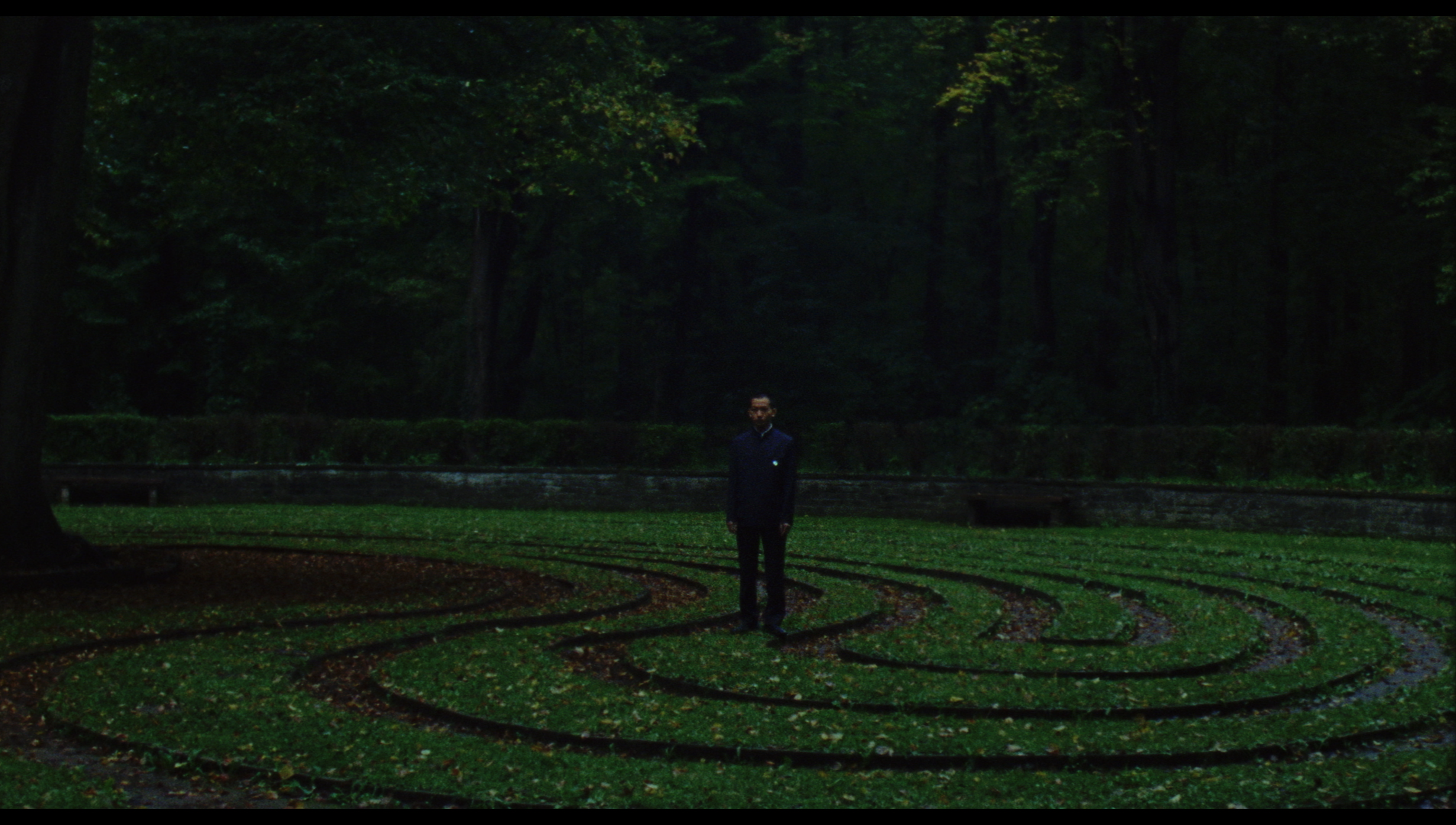 A person stands in the center of a labyrinth made of short grass in a forest. Tall trees surround the area, creating a dense canopy. The scene is dark and moody, with muted lighting and lush greenery.