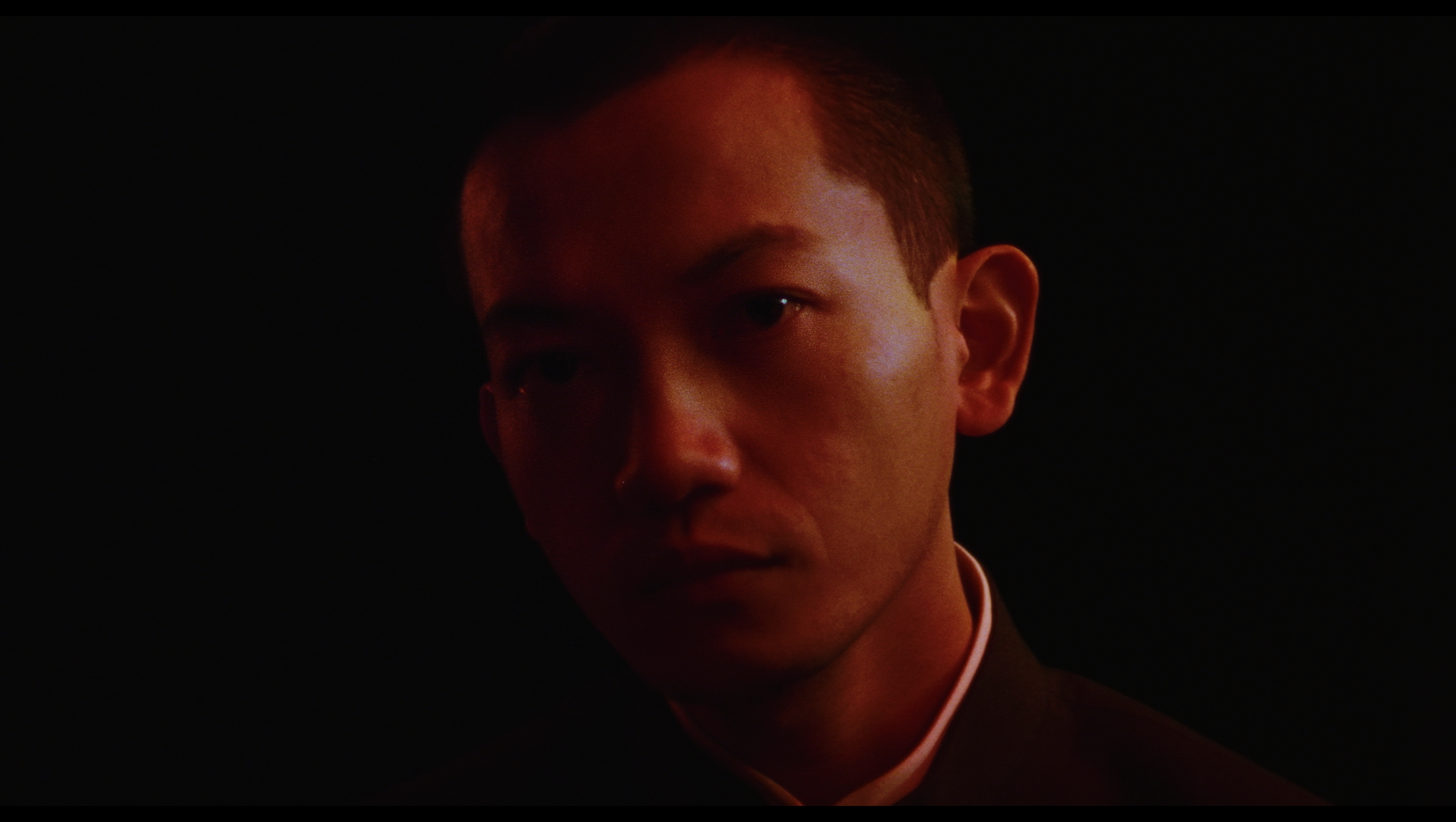 Close-up of a young man with short hair, wearing a dark suit and white shirt, partially illuminated by red light, against a black background.