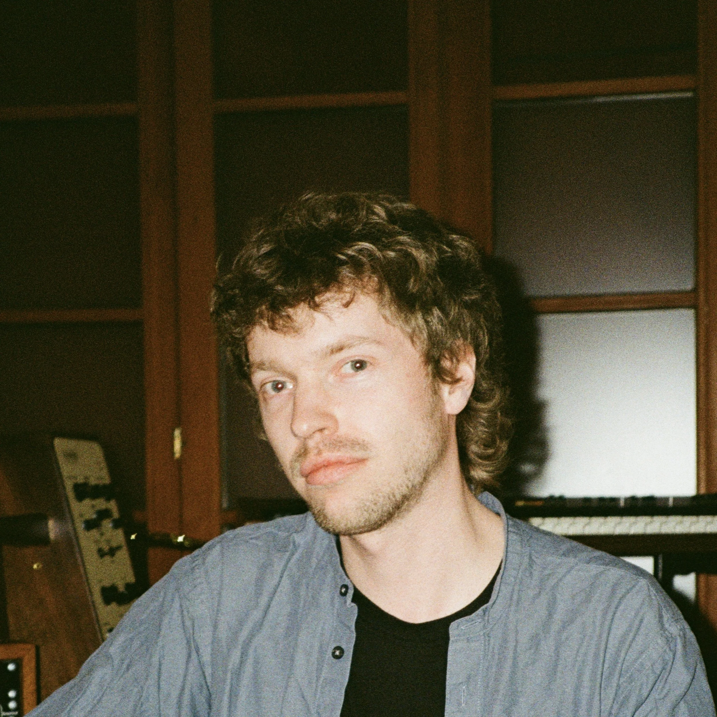 Young man with curly brown hair, light skin, wearing a gray button-up shirt over a black t-shirt, standing indoors in front of a dark wood shelving unit.