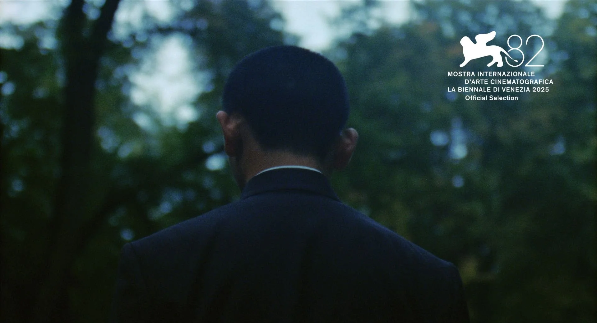 Back view of a man in a suit with short hair, standing outdoors in a forested area with blurred green trees and light in the background. The image includes a logo for the 2025 Venice International Film Festival.