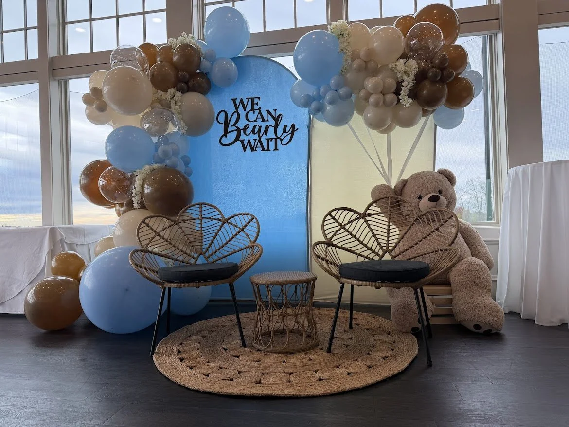 Decorative baby shower setup with a balloon arch, teddy bear, and a banner saying 'We Can Bearly Wait' inside a room with large windows.
