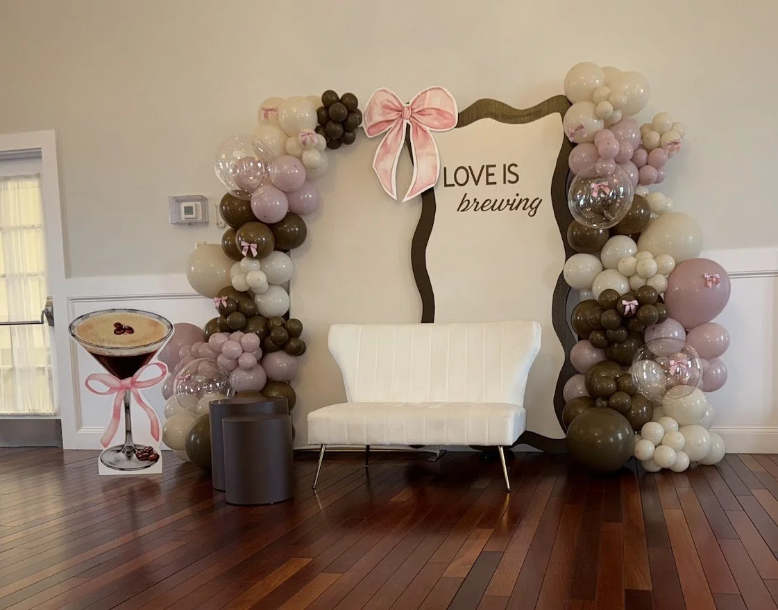 Decorative backdrop with balloon arch in pink, white, brown, and clear balloons, adorned with a pink bow and a label that says "LOVE IS brewing". There is a white sofa, a small gray trash can, and a large cutout of a martini glass with a pink bow and
