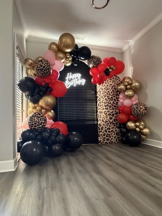 Balloon arch with pink, black, red, gold, and leopard print balloons and a neon sign that says 'Happy Birthday' in a room with wooden floors.