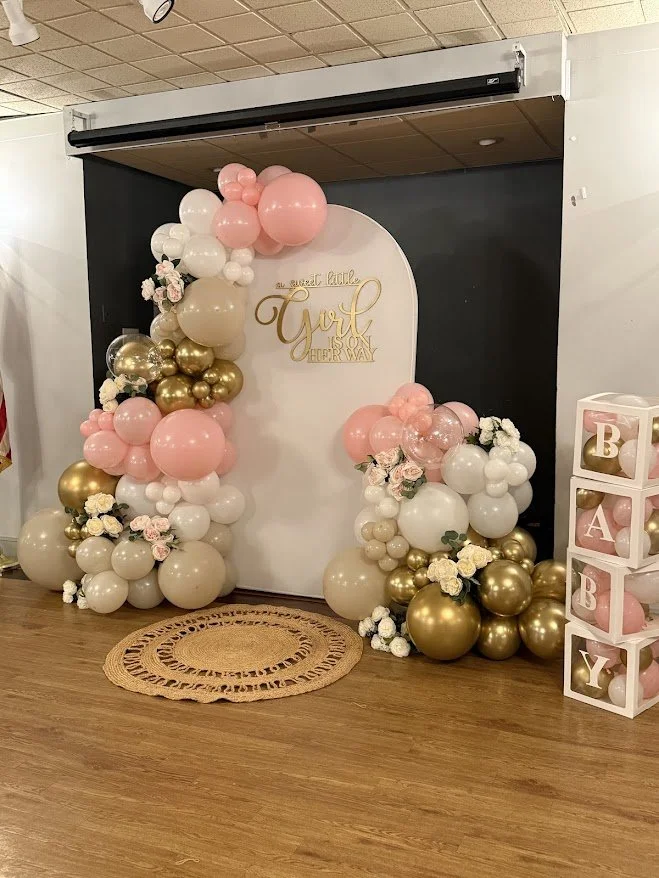 Balloon decoration with pink, white, and gold balloons around a white backdrop and a sign that says "a sweet little girl is on her way"; pink and white flowers at the base; a bubble letter "BABY" block on the right.