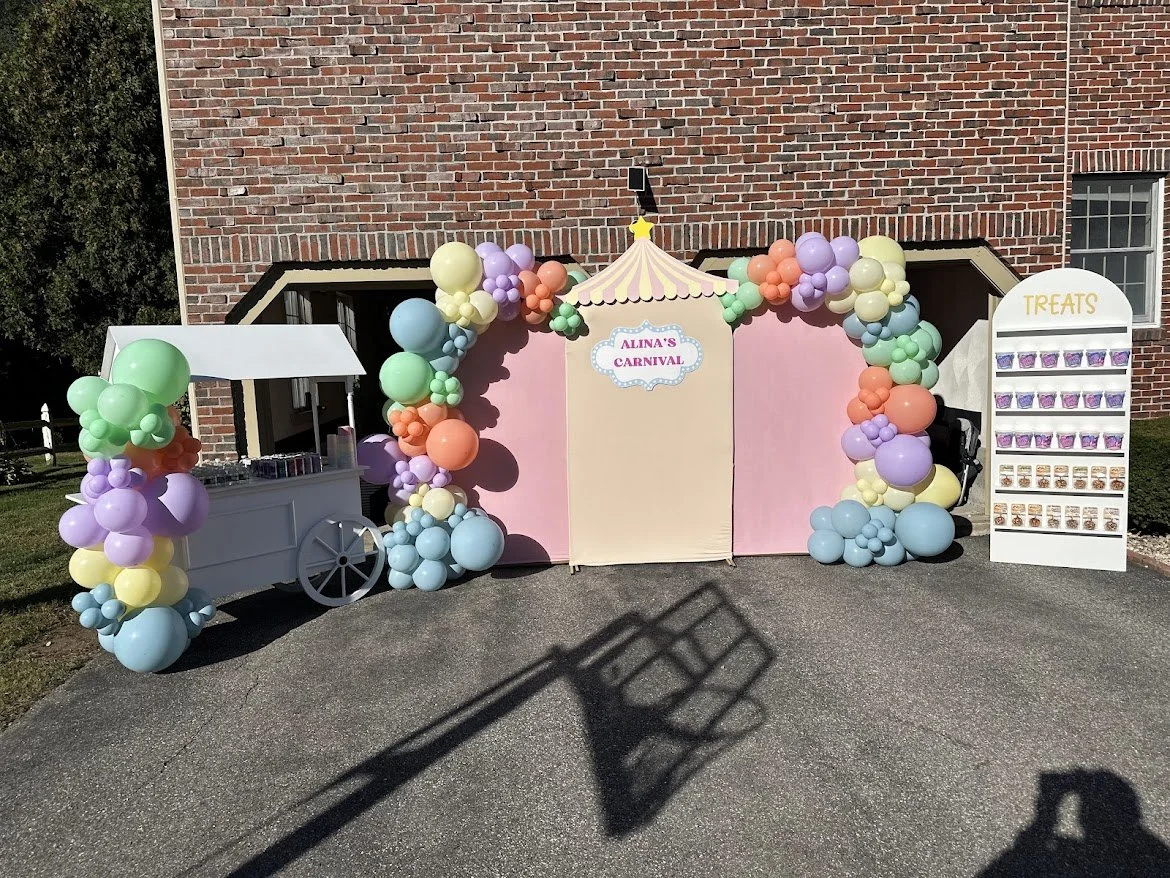 Decorative carnival-themed photo booth with balloon arch, a pink backdrop, and a treat stand labeled "Treats" with candy jars. A sign reads "Alina's Carnival."