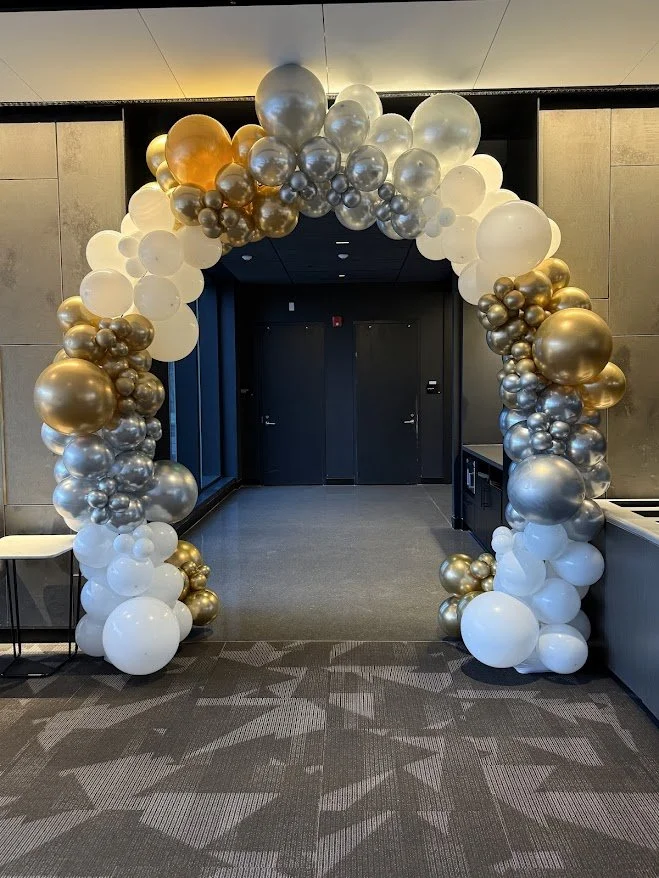 Balloon arch decorated with white, gold, and silver balloons at an indoor event entrance.