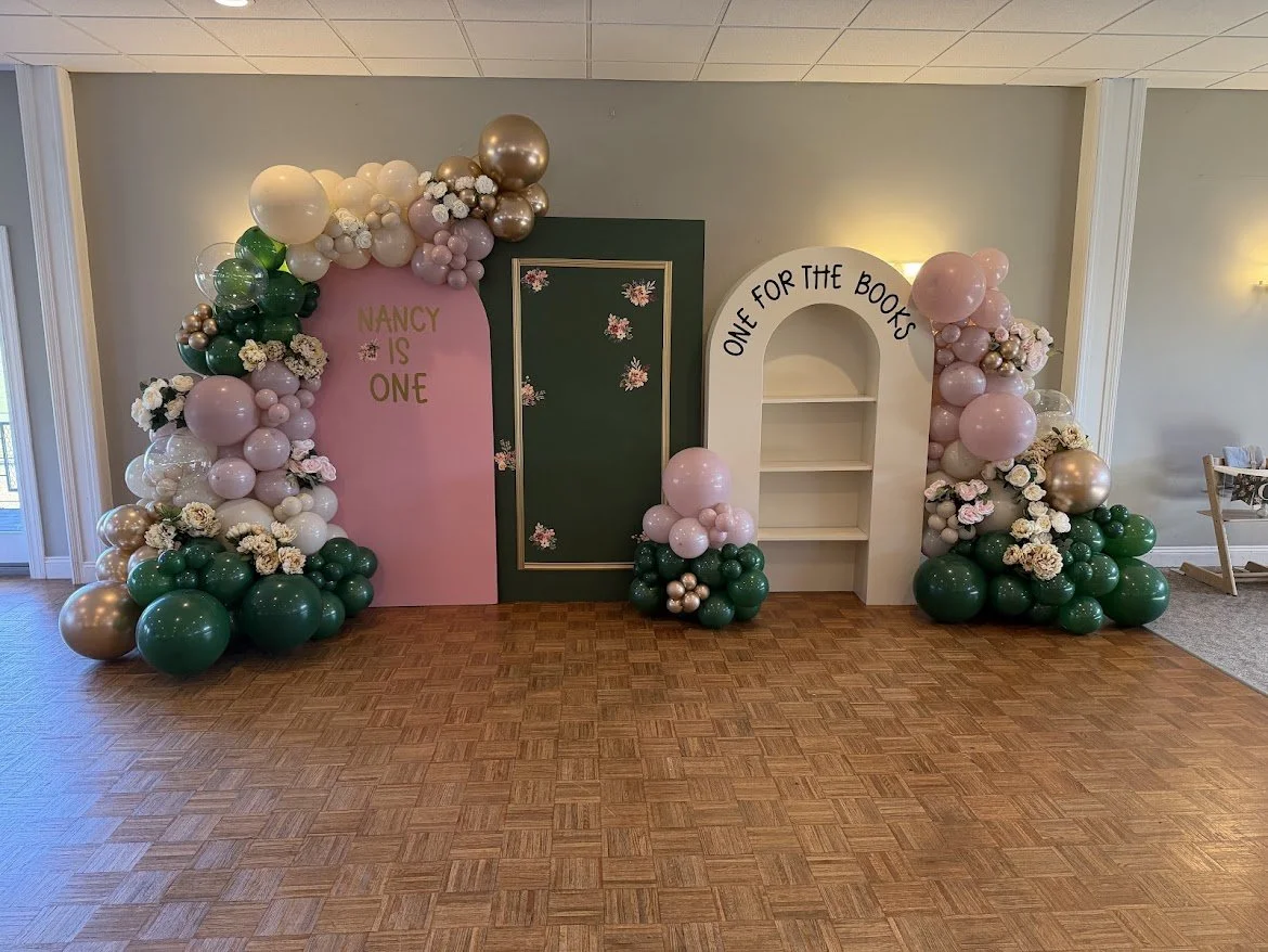 Decorative party backdrop with balloon arrangements, pink, green, white, and gold balloons, floral accents, and a shelving unit. One side has a pink sign with 'Nancy is One', and the other side has 'One for the Books'.