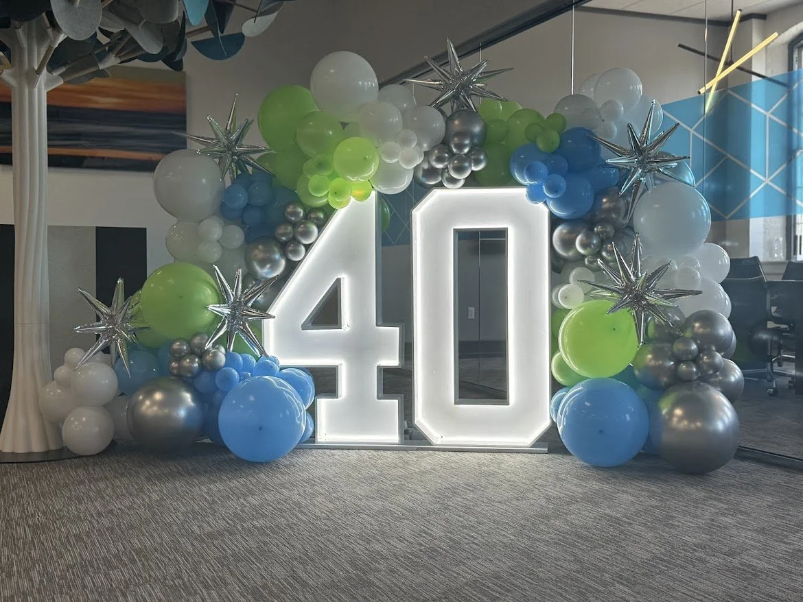 Large illuminated number 40 surrounded by colorful balloons and metallic star-shaped decorations, likely for a celebration or milestone event.